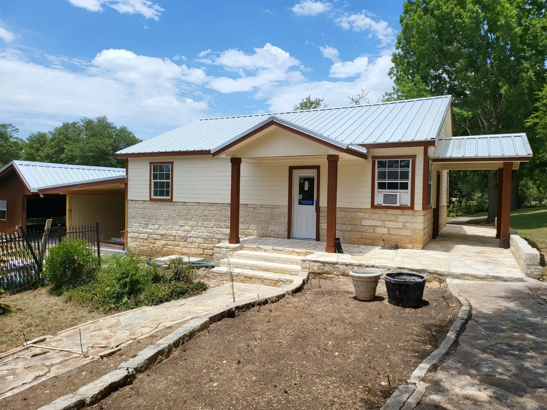 A house under construction with a metal roof, beige siding, and stone foundation, featuring a front porch with stairs, surrounded by a mostly unfinished yard with some plants and construction materials.
