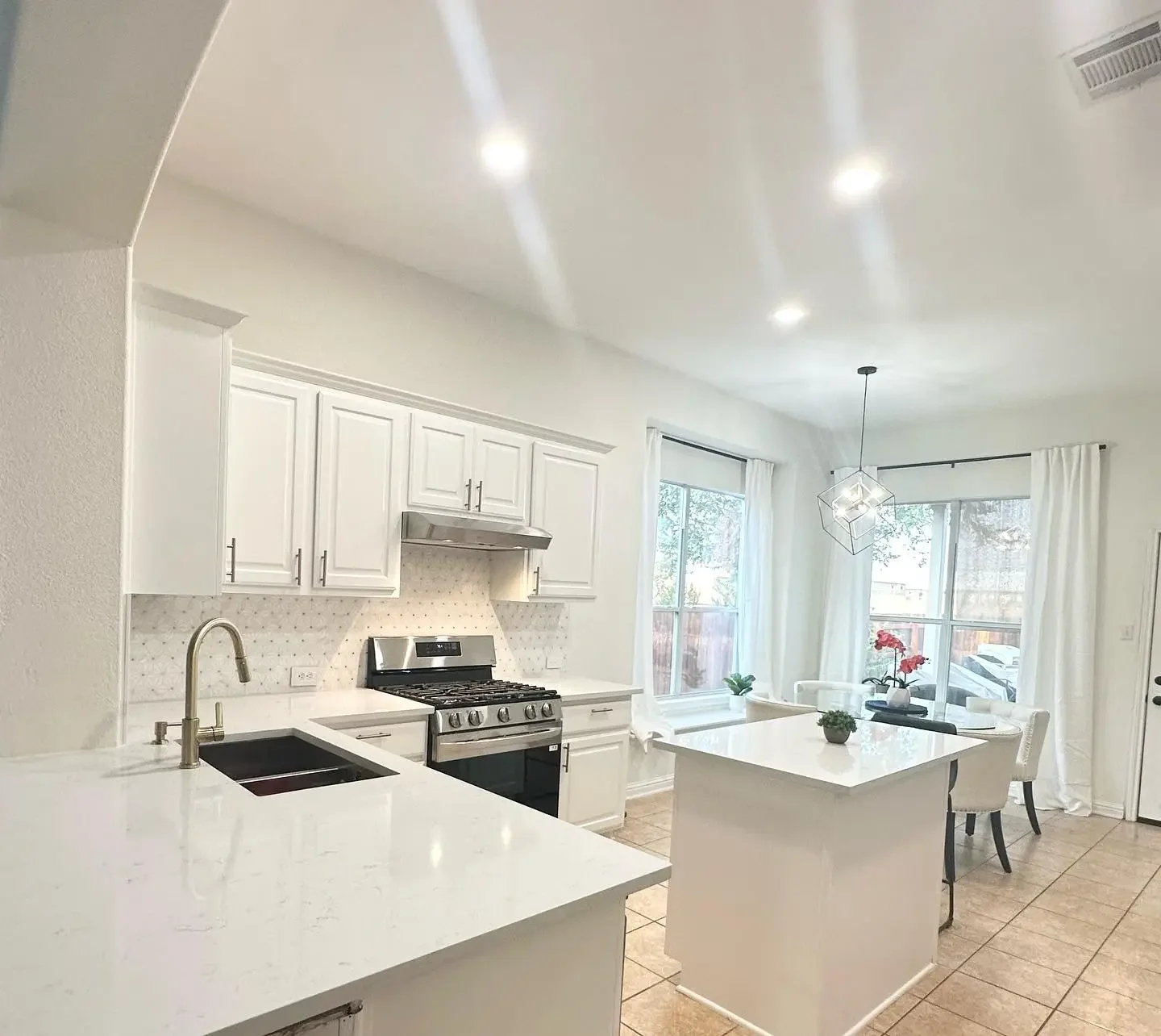 Bright kitchen with white cabinets, stainless steel stove, white countertops, small dining area with four chairs around a white table, large windows with white curtains, tiled floor, and a geometric pendant light.