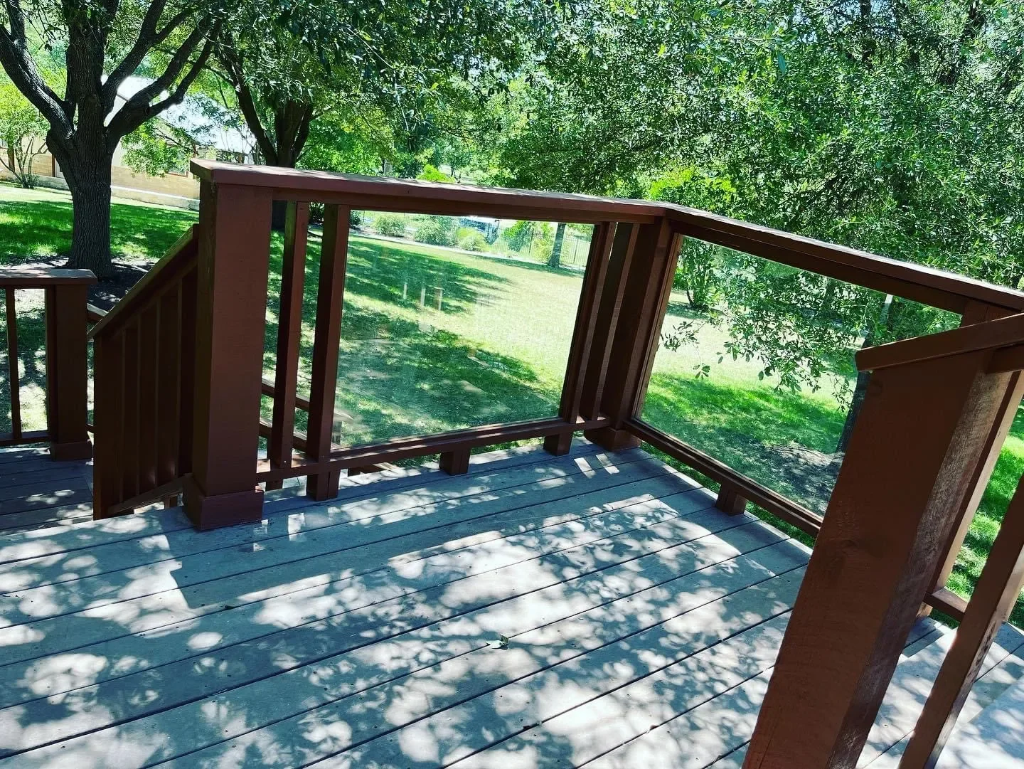 View of a wooden deck with safety railing, surrounded by green trees and grass, with shadows of leaves on the deck.