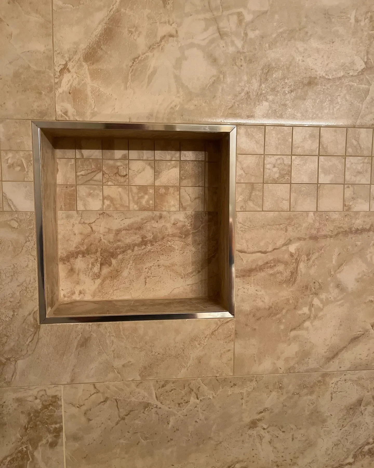 A square, recessed shower nook framed with brushed metal, set into a beige, marbled tile wall with a strip of smaller square tiles.