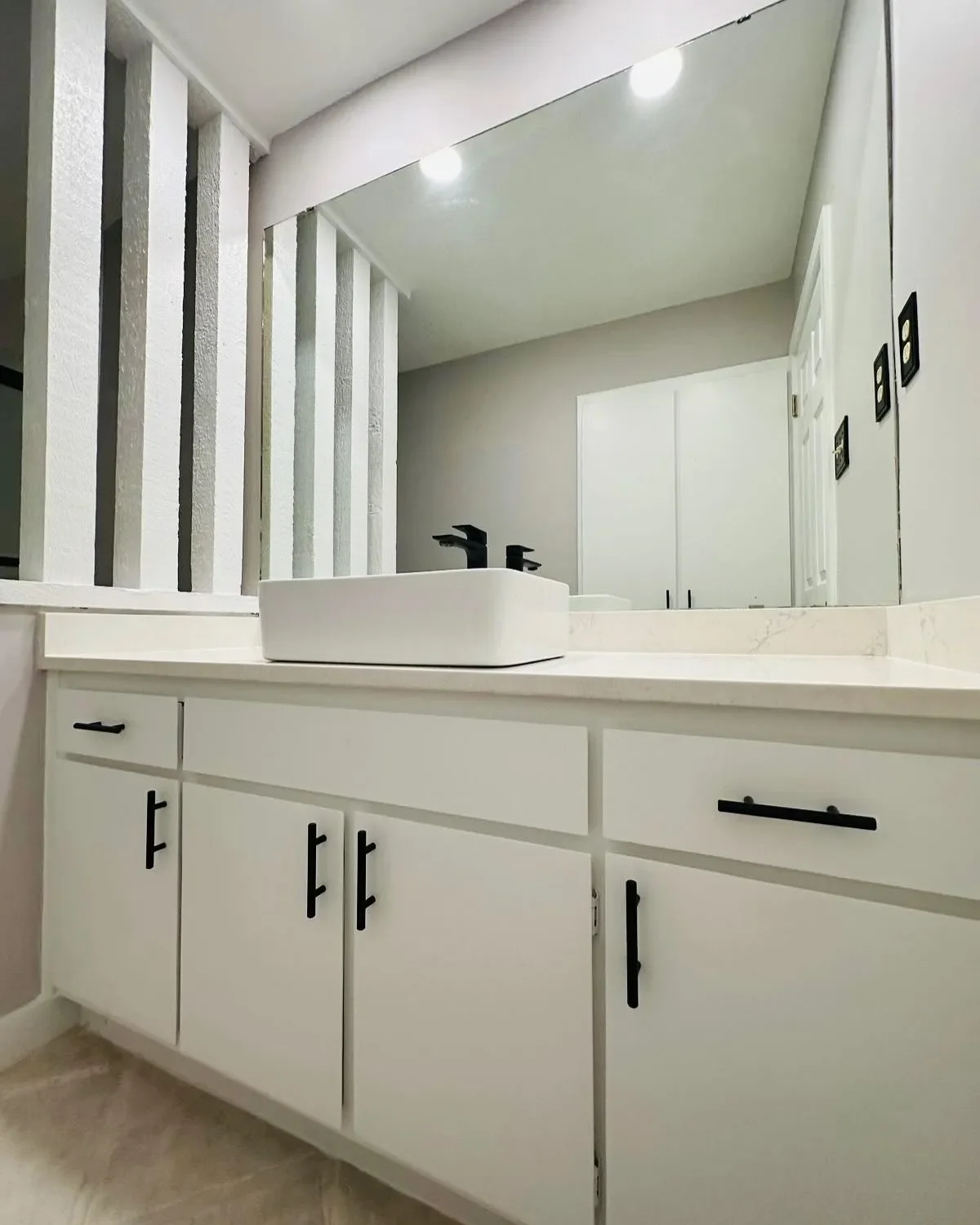 A modern bathroom vanity with a white cabinet, black handles, a white vessel sink, a black faucet, and a large mirror above.