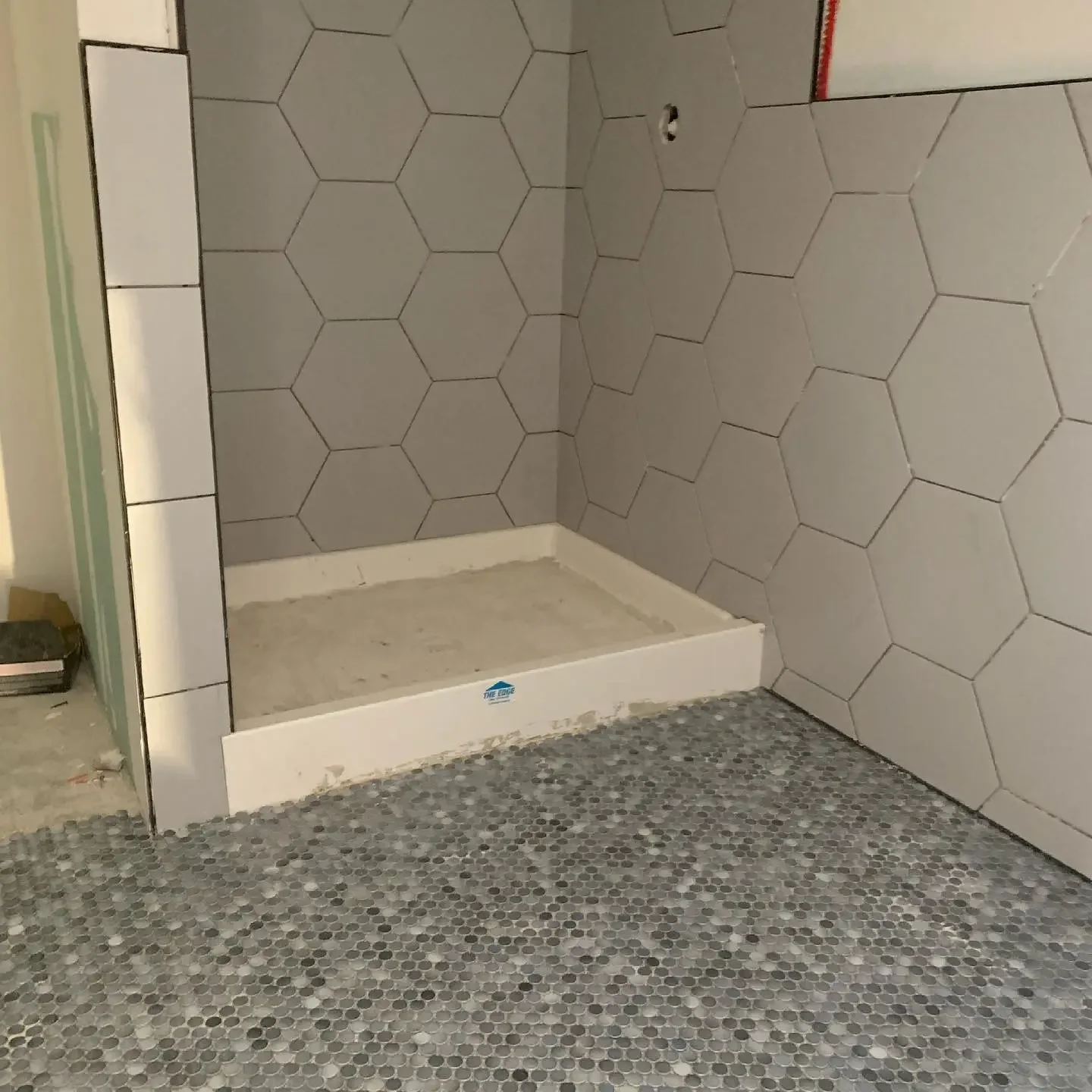 A bathroom or shower area under construction with hexagonal wall tiles and small mosaic floor tiles, and a shower base installed.