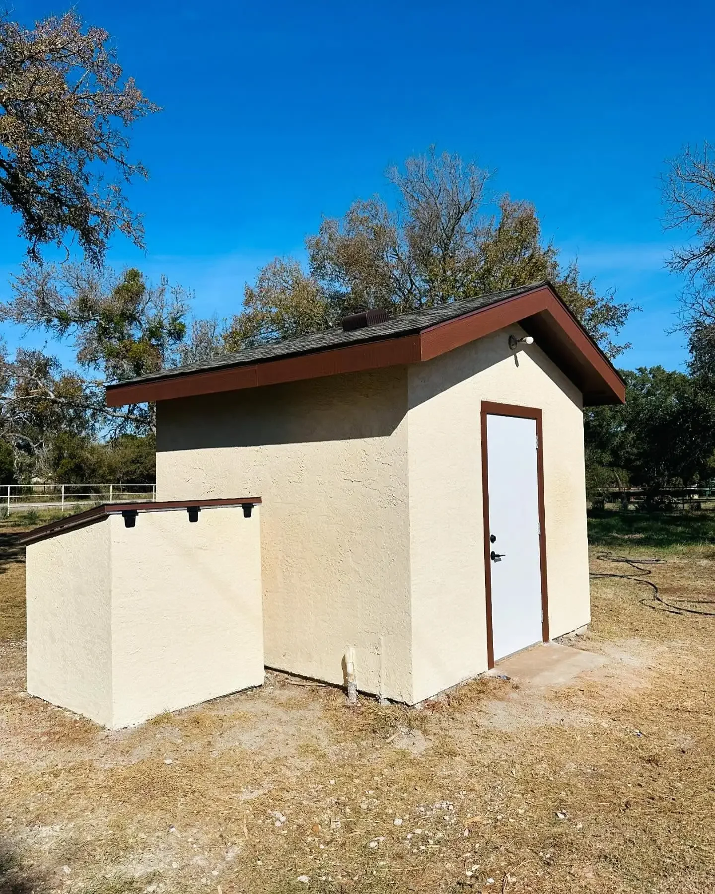 Small beige building with a white door, brown trim, and a sloped roof, set outdoors on a dirt ground with trees and a blue sky in the background.