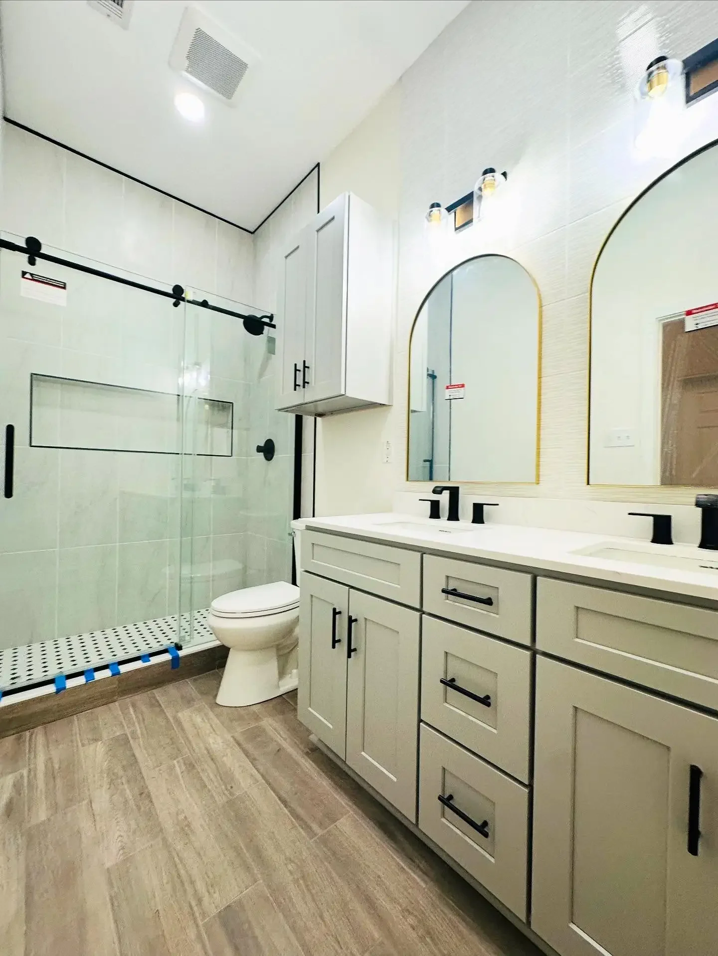 Modern bathroom with double vanity sinks, large mirrors, soft beige cabinets, black fixtures, and a glass-enclosed shower with black hardware and wood-look tile flooring.