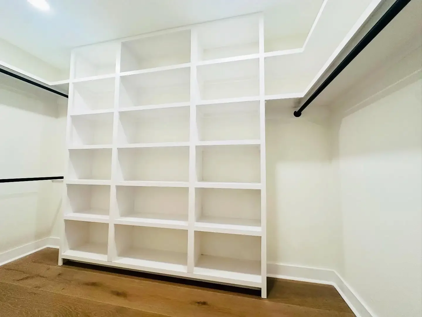 Empty walk-in closet with white shelving unit and black hanging rods on both sides, hardwood floor, and white walls.
