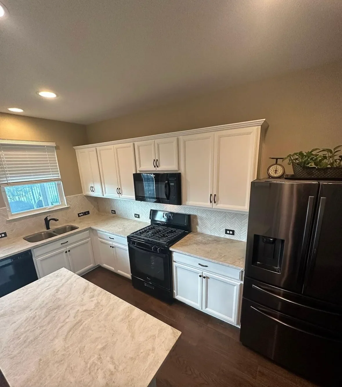 Modern kitchen with white cabinets, black appliances, beige countertops, wood flooring, and a small window with blinds