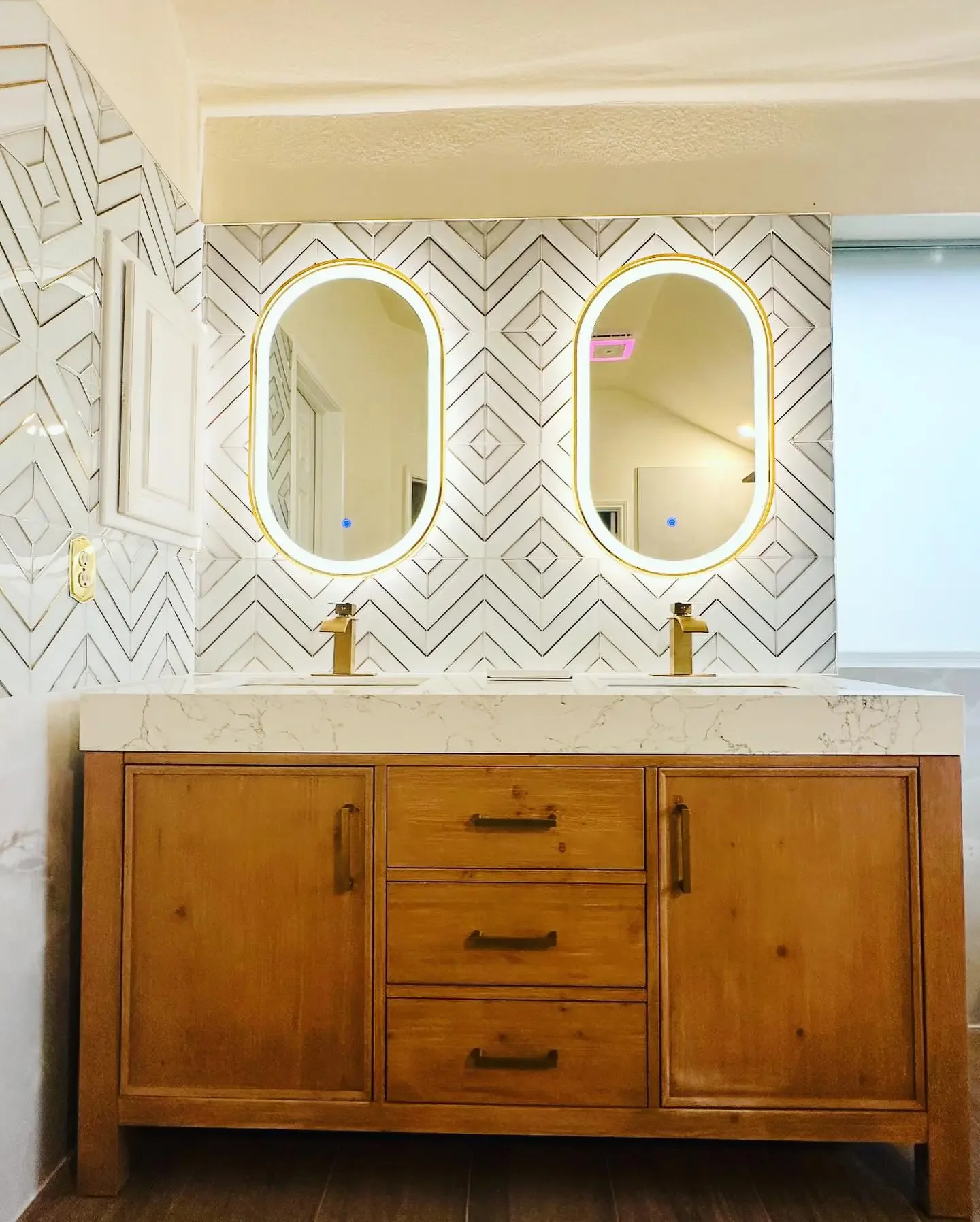 Modern bathroom vanity with double oval mirrors backlit with LED, wood cabinet with three drawers and two doors, marble countertop, and geometric patterned tile wall.