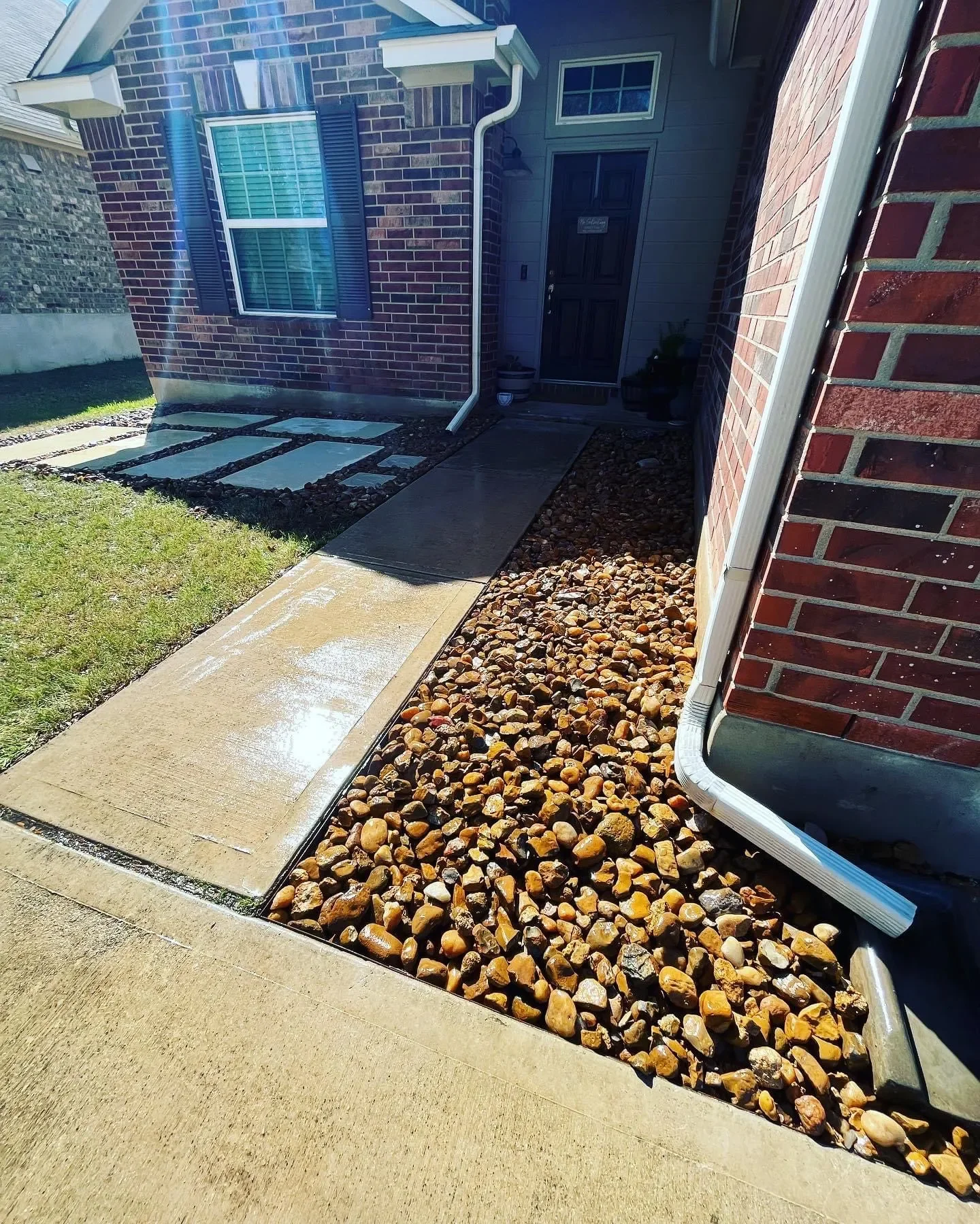 Front step and pathway leading to a house entrance, with a concrete walkway, decorative rocks alongside, brick siding walls, and a front door in the background.