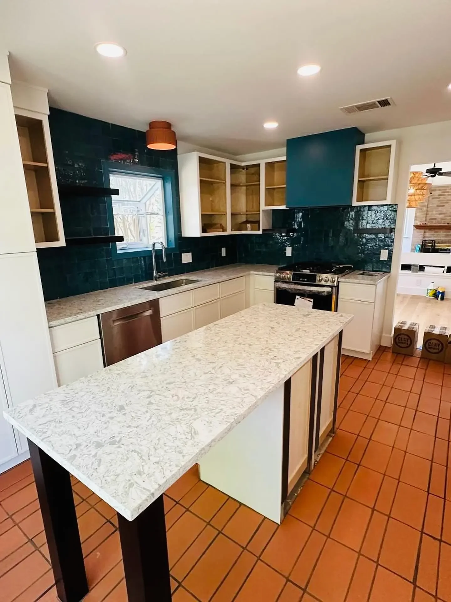 Kitchen with white cabinets, a granite countertop, a small island, open shelves, green tiled backsplash, and terra cotta floor tiles.