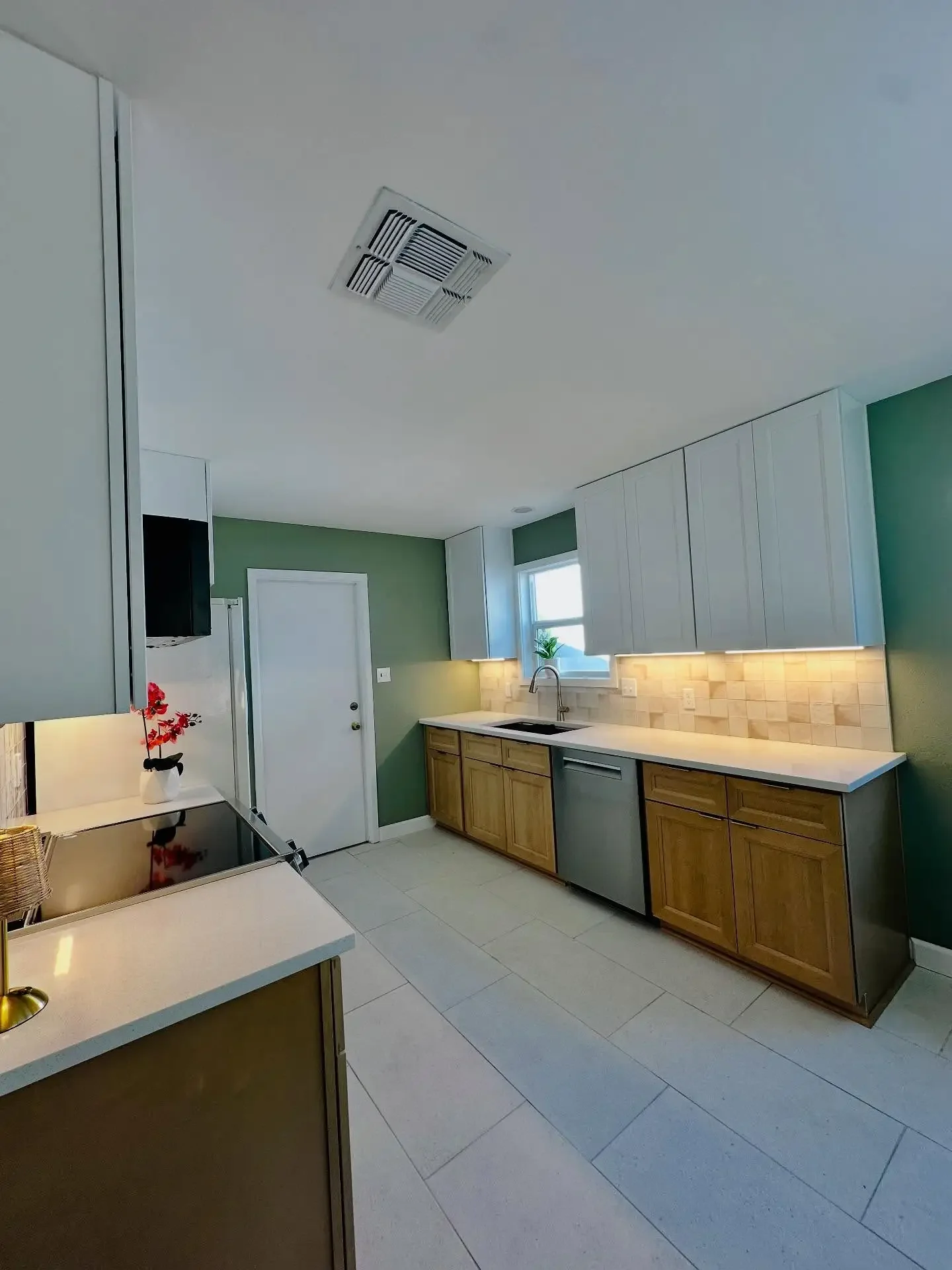 Modern kitchen with green walls, white cabinets, wood lower cabinets, beige backsplash, stainless steel dishwasher, black cooktop, and small plants on the windowsill.