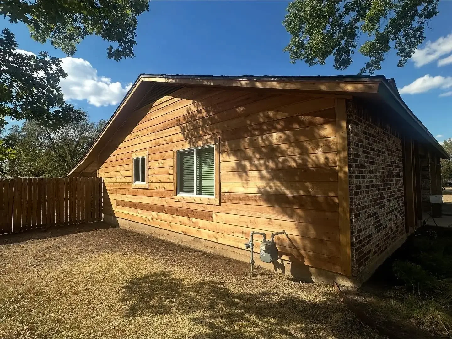 Rear view of a house with new wooden siding and a brick section, two small windows, a wooden fence, and landscaping under a sunny sky with trees.