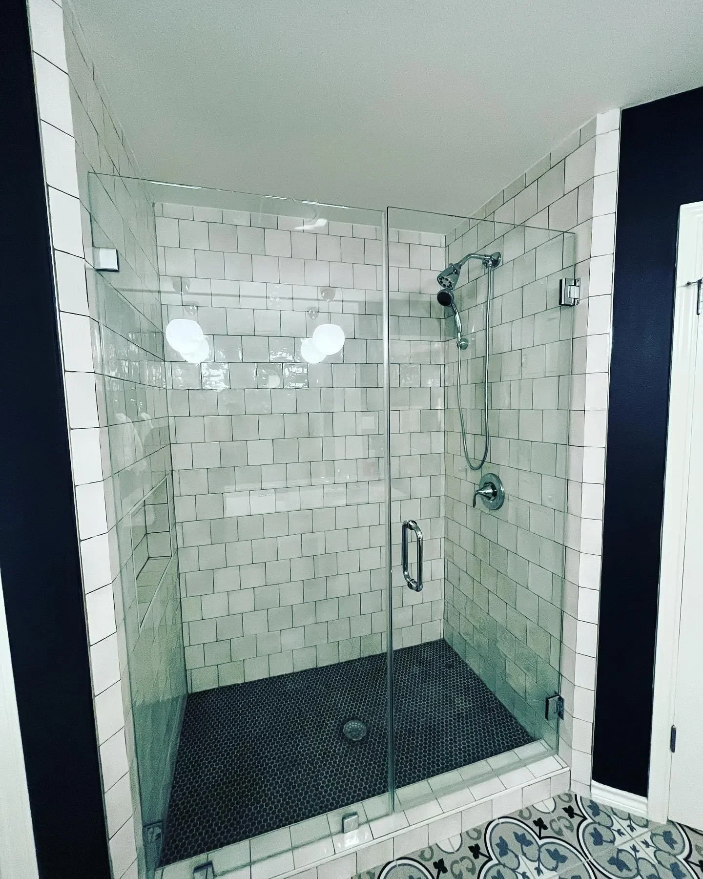 Glass shower enclosure with white subway tiles, black pebble floor, adjustable showerhead, built-in wall niche, and geometric patterned tile floor outside.