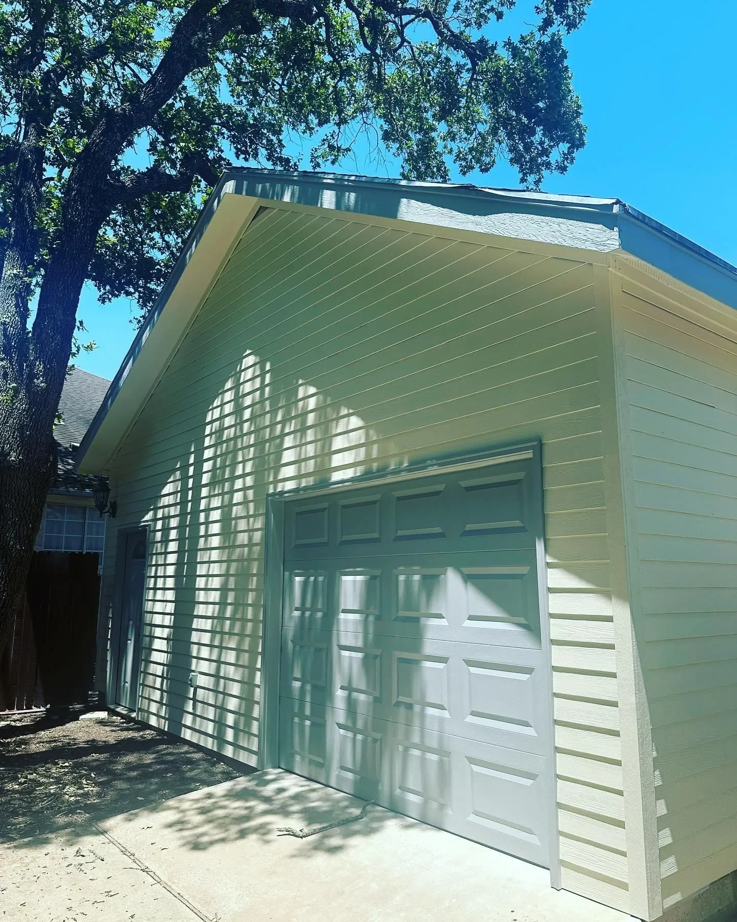 A light-colored house garage with a closed garage door, shadow of tree branches cast on the wall, next to a large tree with green leaves, under a clear blue sky.