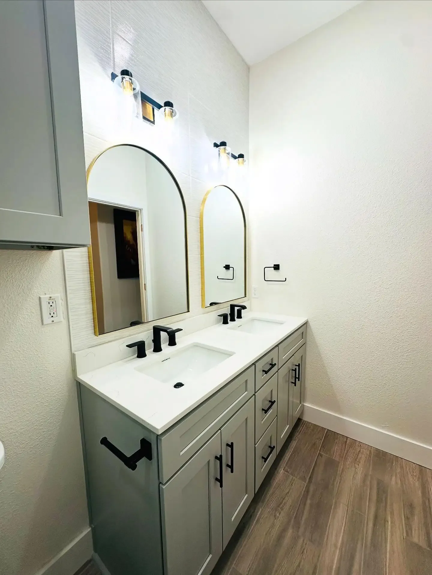 Modern bathroom vanity with dual sinks, gray cabinets, black fixtures, two large mirrors, and wooden flooring.