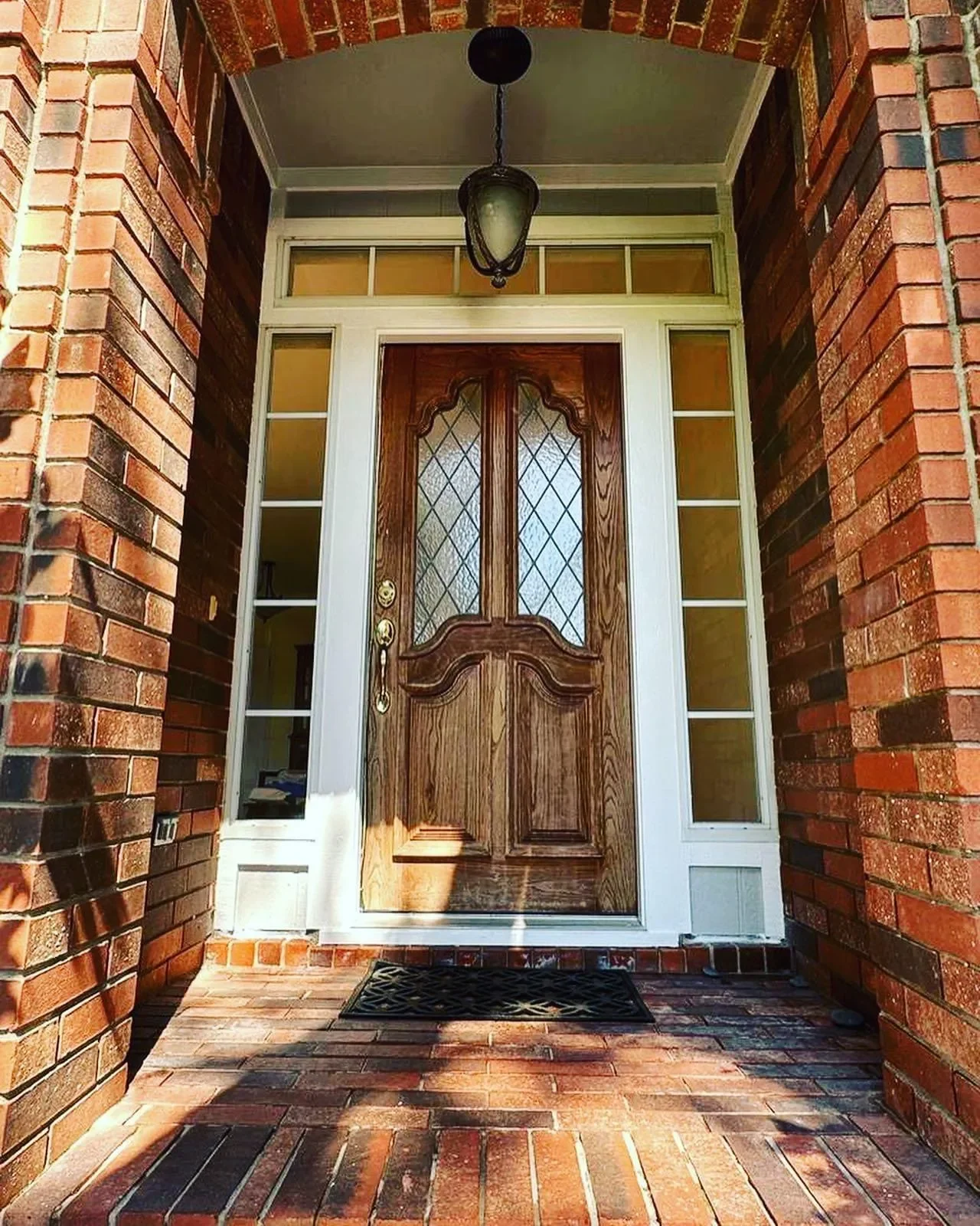 Front door with wooden panels and glass windows framed by white trim on a brick porch with an overhead lantern.