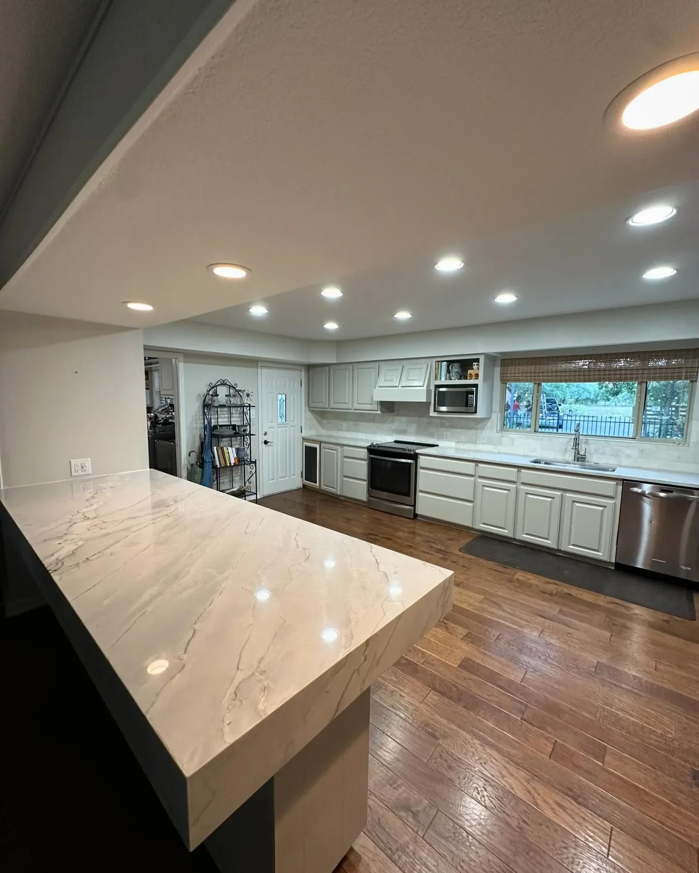 Modern kitchen with white cabinets, stainless steel appliances, hardwood floors, and a large window over the sink, illuminated by recessed ceiling lights.