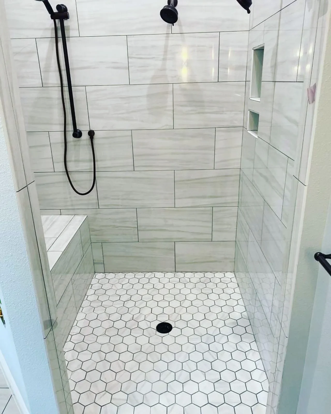 A walk-in shower with large white rectangular wall tiles, a built-in shelf, a bench, and white hexagon floor tiles with a drain in the center.