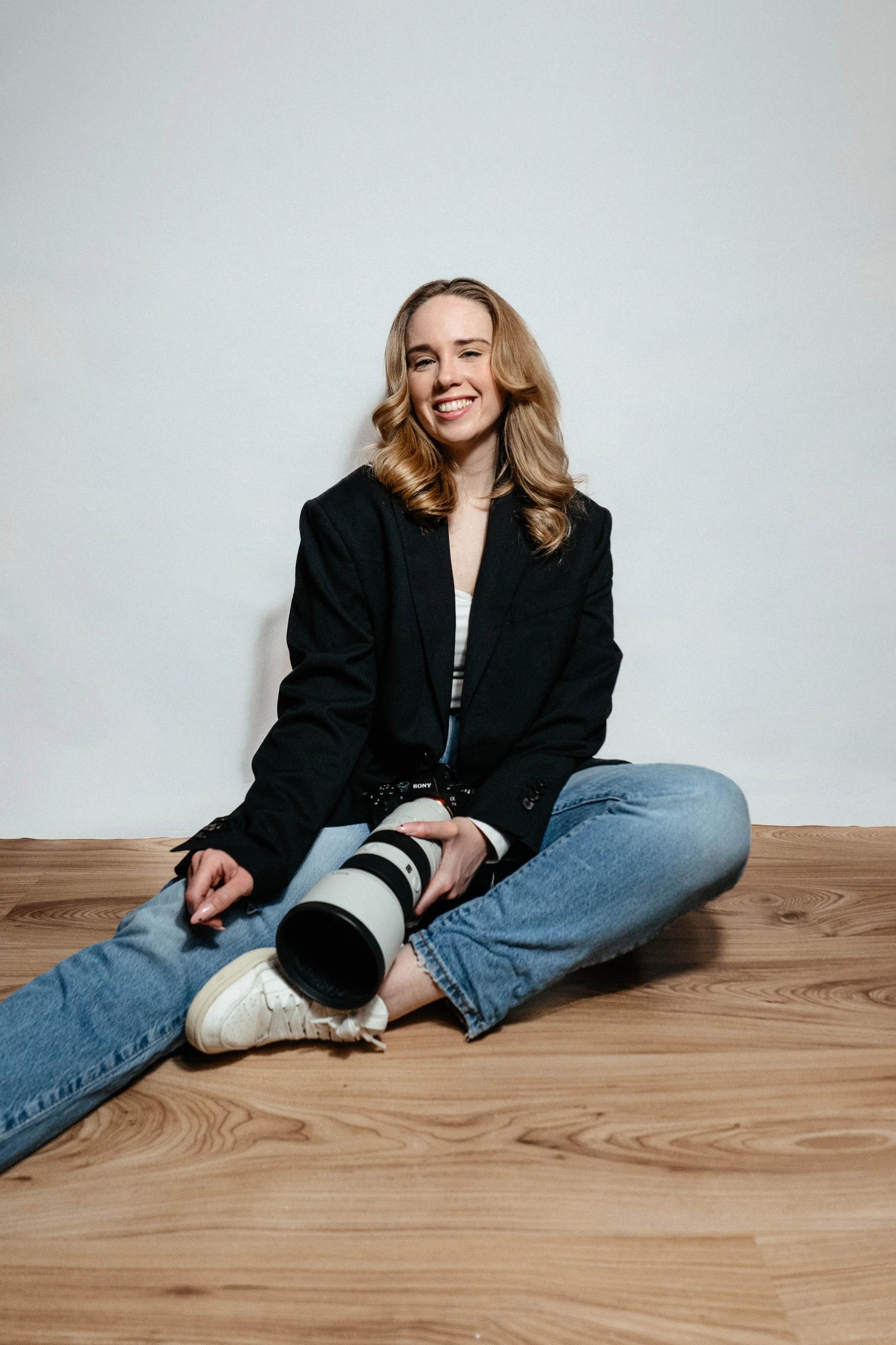 A woman sitting on a wooden floor smiling, holding a professional camera lens, wearing a black blazer, striped shirt, jeans, and white sneakers, with a plain white background.