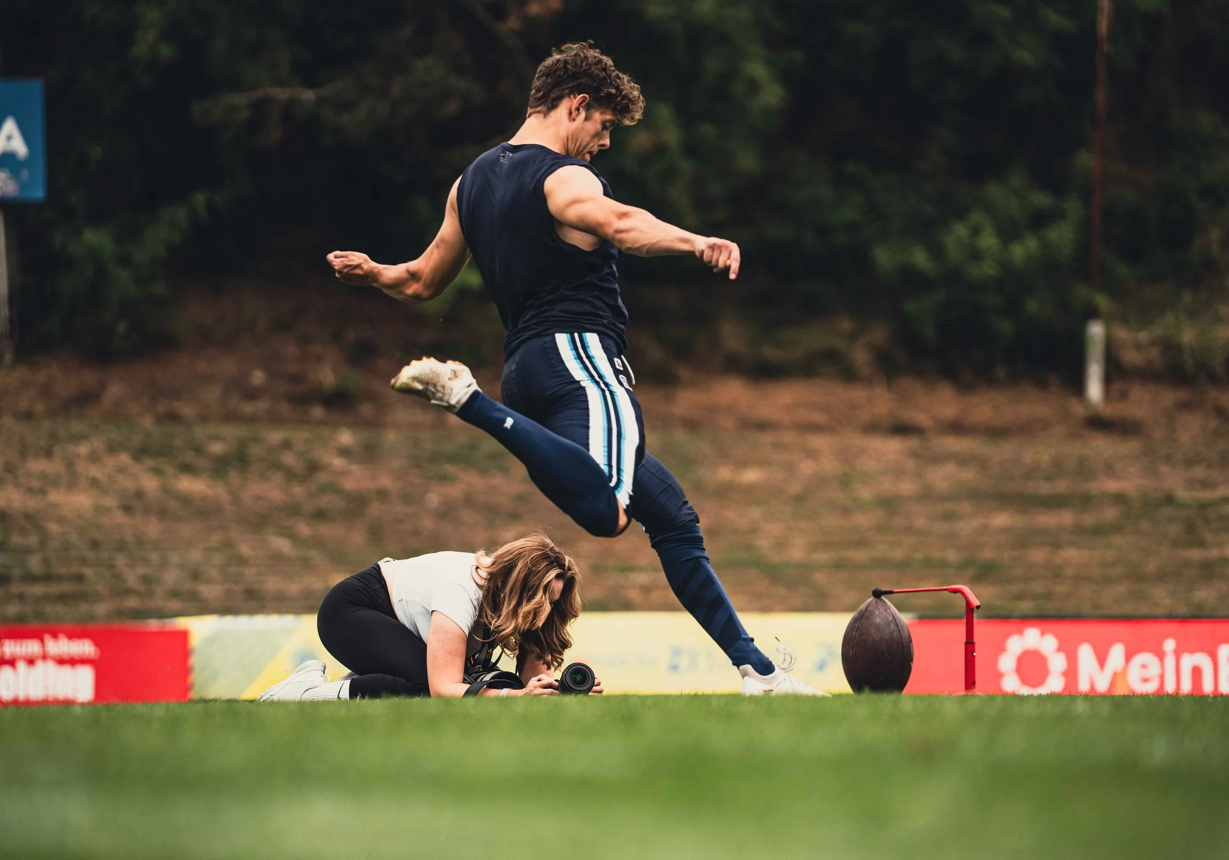 A man jumping over a woman who is lying on the ground and taking a photo, on a sports field with a blurred background and a red banner.