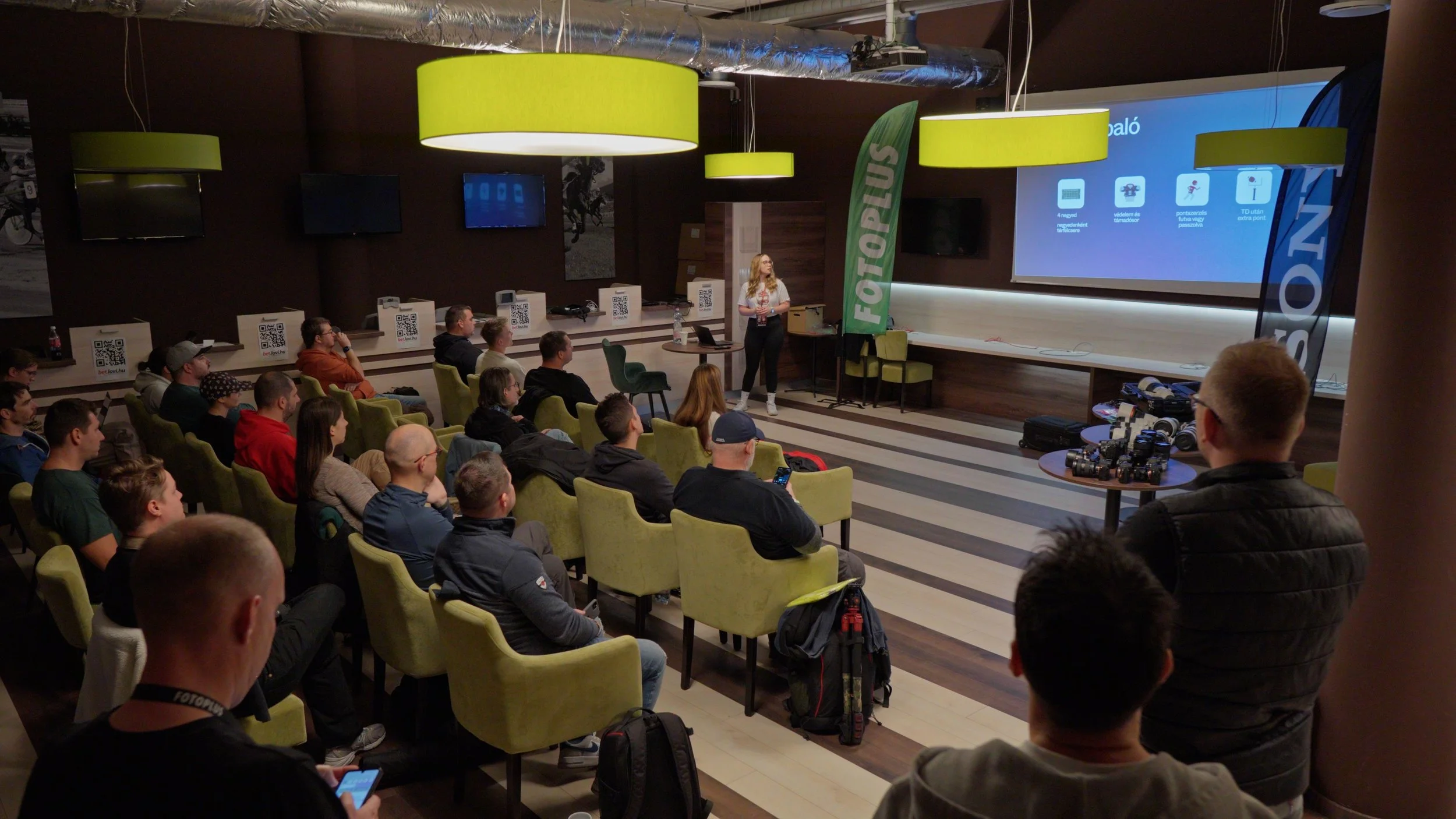 A woman stands in front of an audience giving a presentation in a conference room. The screen behind her displays information about a product or service, and there are banners with the word 'FOTOPLUS'. The audience is seated in green chairs, some are taking notes or photos, and there are tables with camera equipment on the right side.