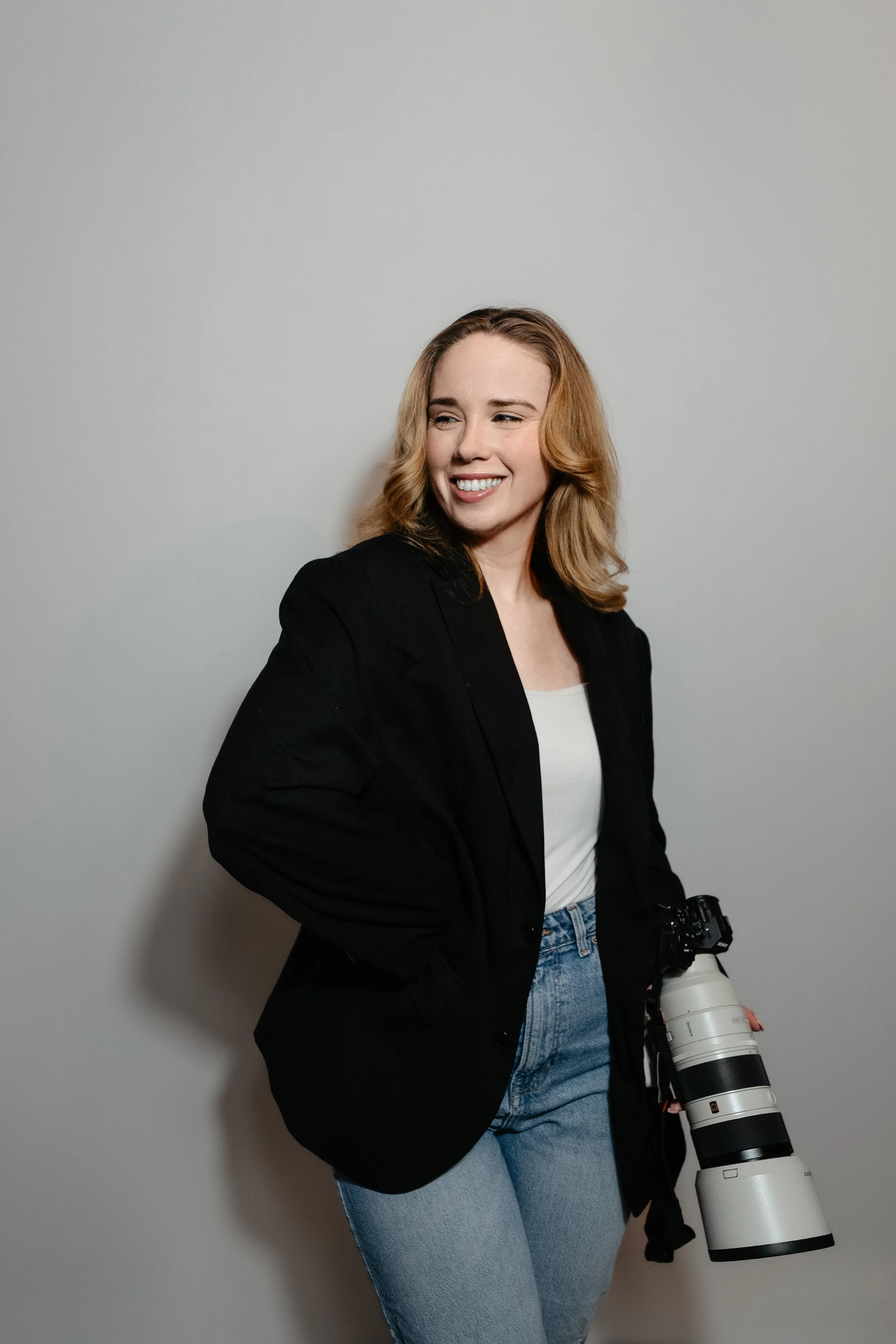 A woman with blonde hair wearing a black blazer and jeans, holding a professional camera with a large telephoto lens, standing against a plain gray background and smiling.