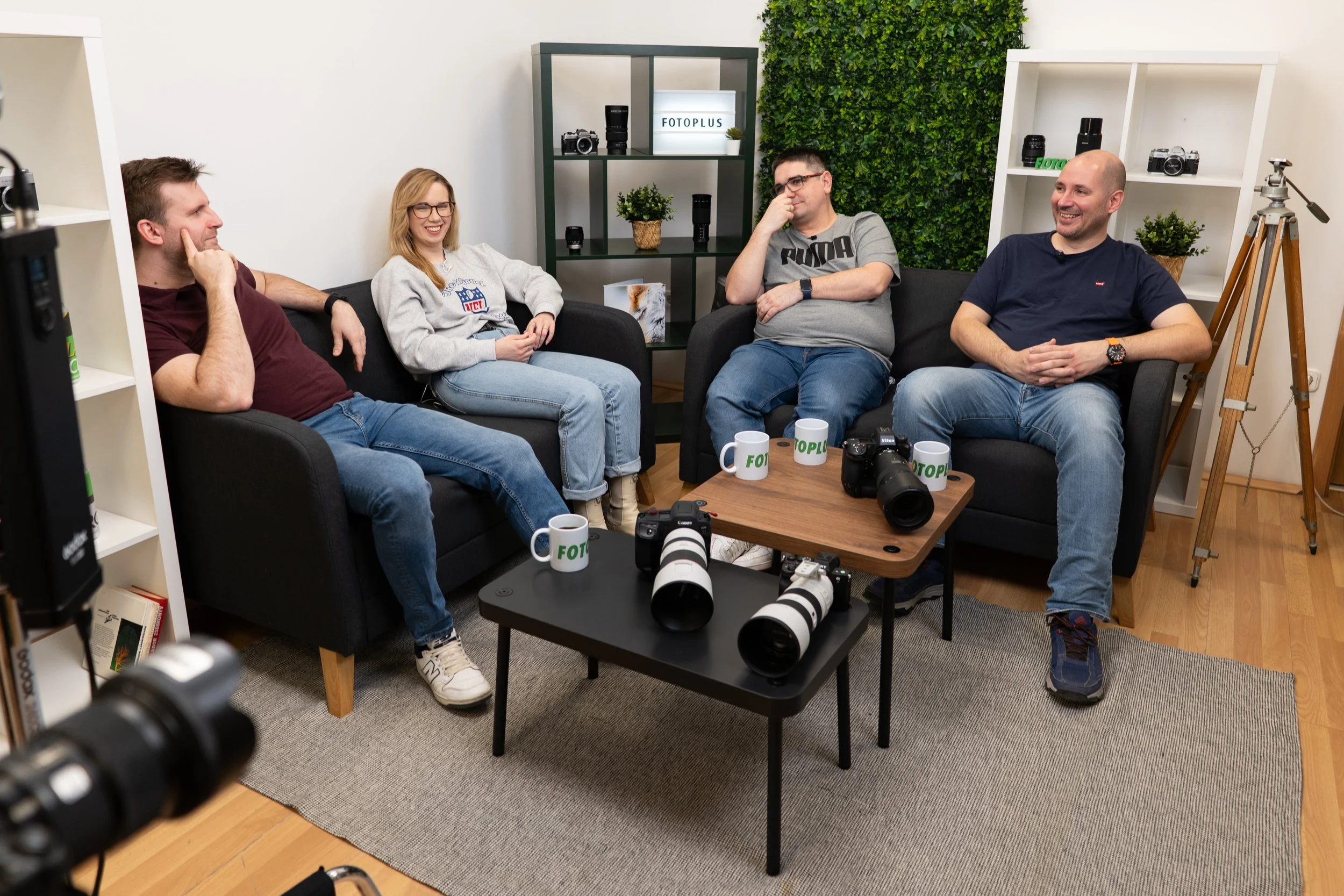 Four people sitting on black sofas in a room with camera equipment and mugs on two tables, engaging in a conversation, with cameras on tripods and a green wall background.