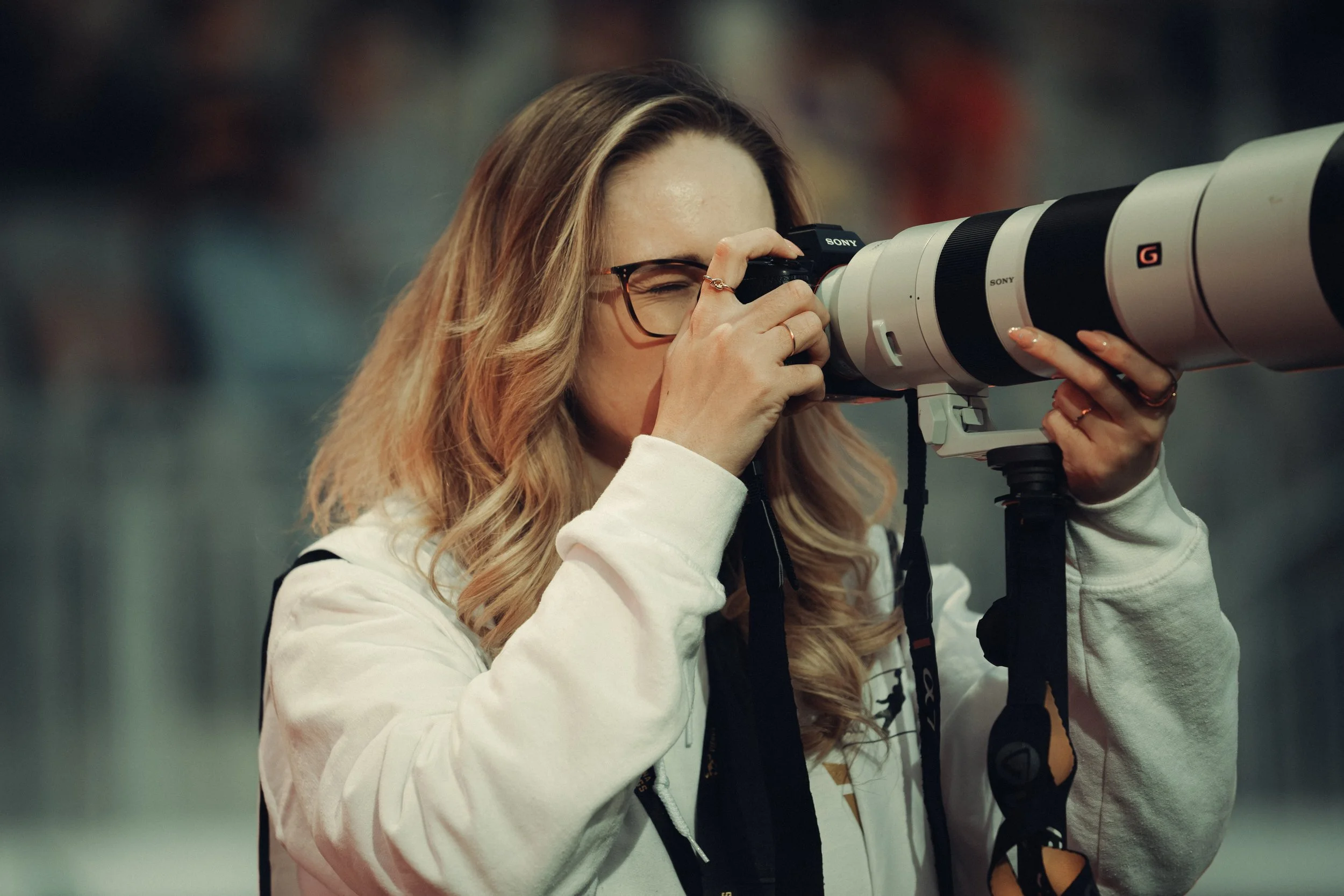 A woman with blonde hair and glasses looks through a large telephoto camera lens.