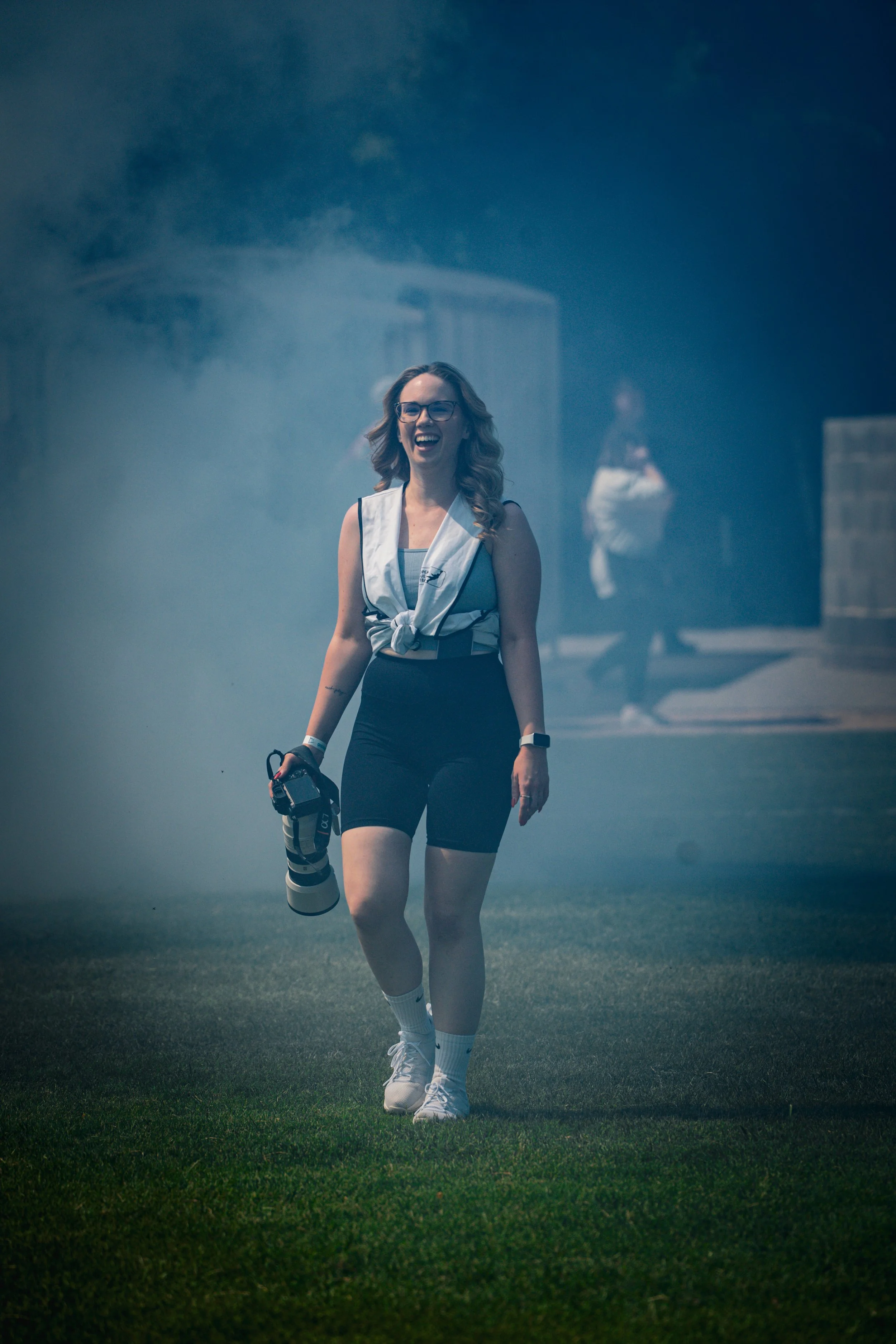A woman with glasses and wavy hair is walking on a grassy field, smiling and holding a camera lens. She is wearing a sleeveless top, black shorts, and white sneakers, with a white jacket tied around her shoulders. In the background, there is another woman and a foggy or smoky atmosphere.
