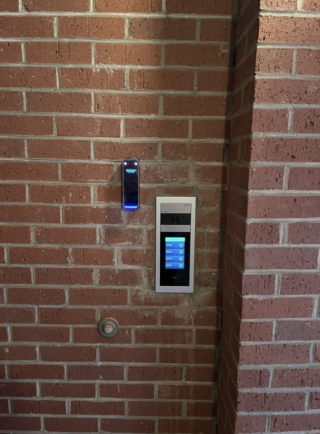 A brick wall with a digital access control panel, a small device with a blue light, and a button installed on it.
