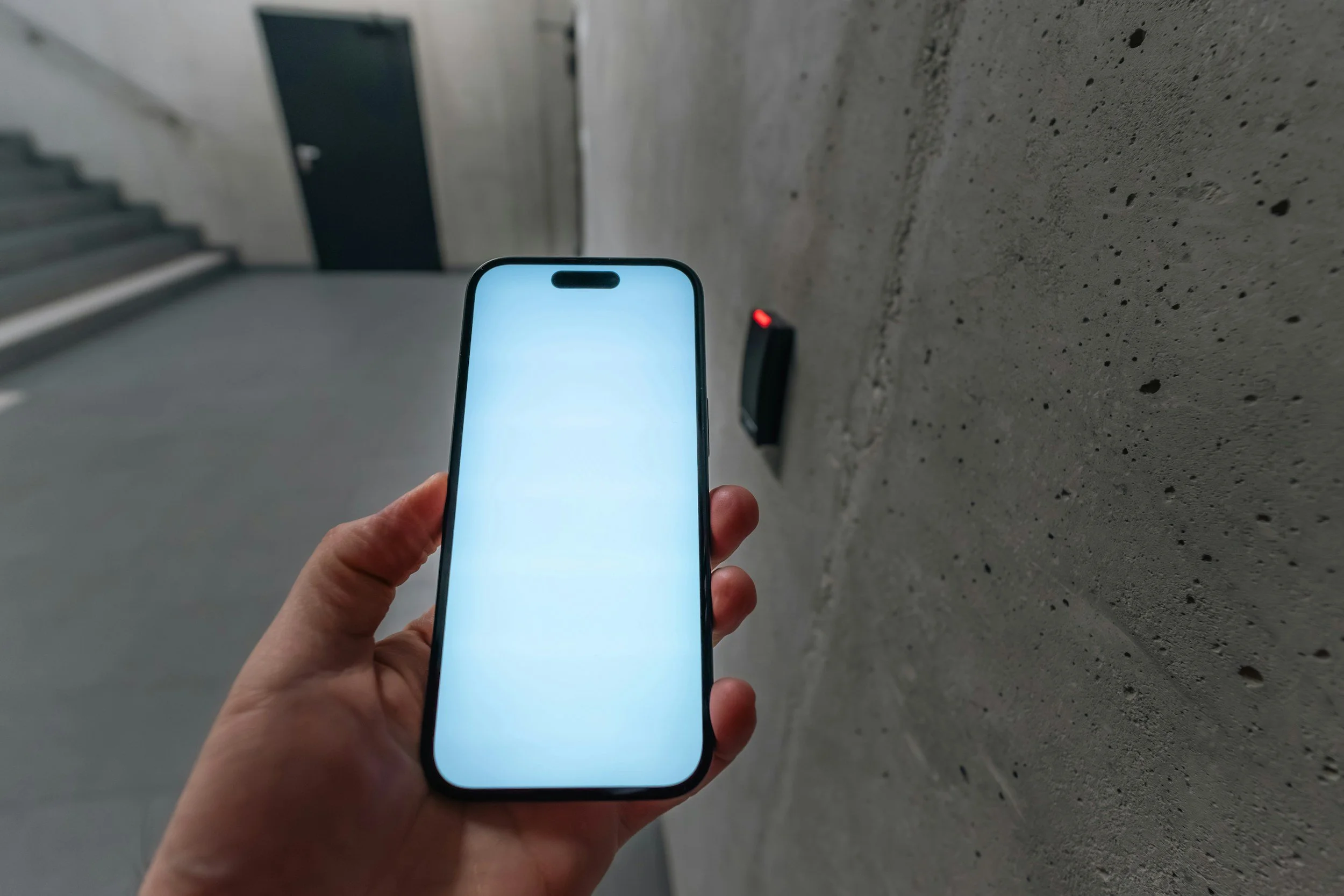 A person holding a smartphone in front of a concrete wall. In the background, there are stairs and a black door. The smartphone screen is turned on and glowing brightly.