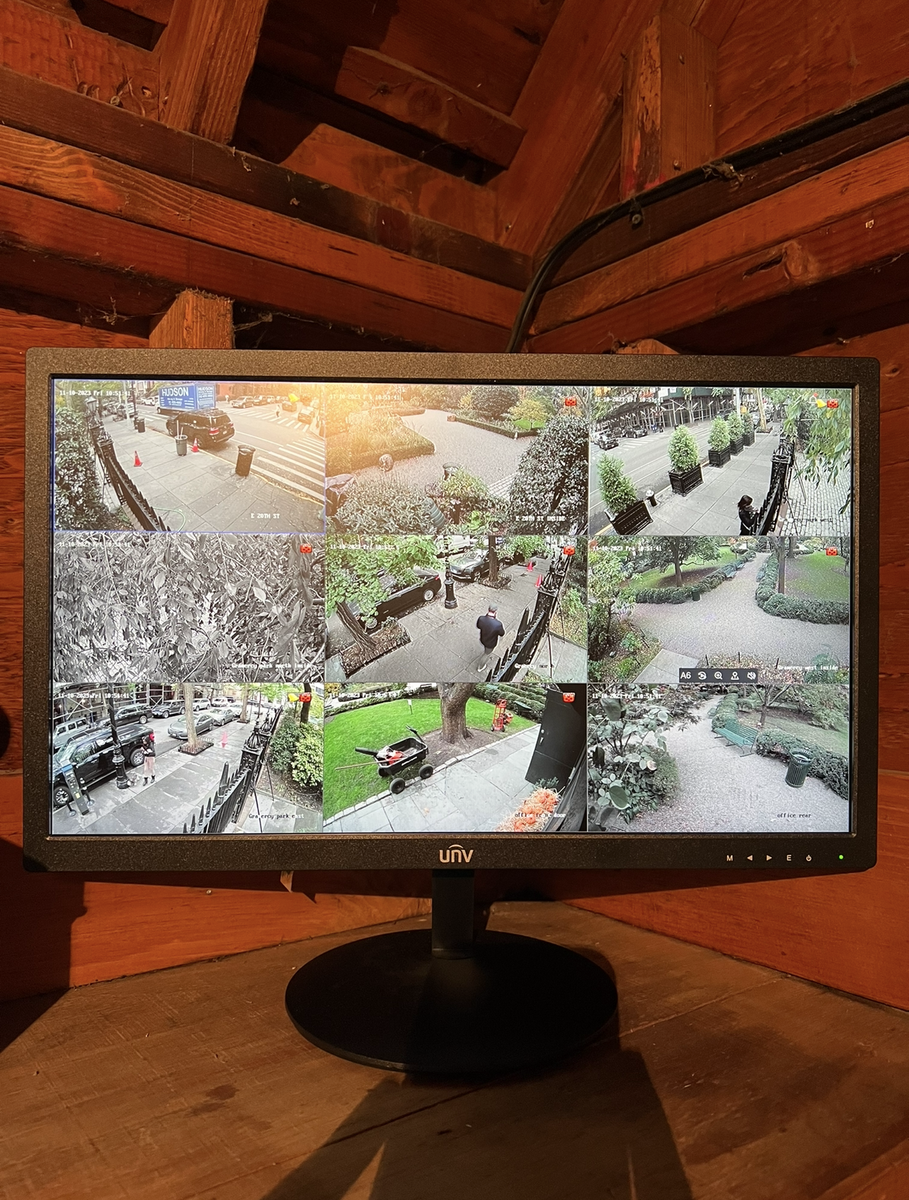 A security monitor displaying nine surveillance camera feeds of outdoor scenes in a city or park, showing trees, benches, pedestrians, and parked cars.