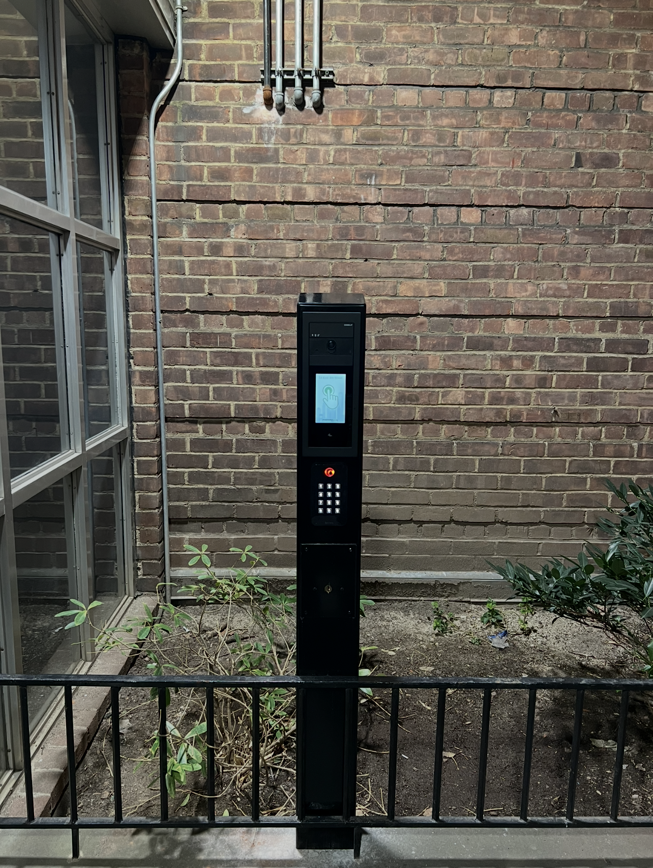 A black intercom station with a touchscreen display, keypad, and camera, situated outside against a brick wall, surrounded by small bushes and a metal fence in the foreground.