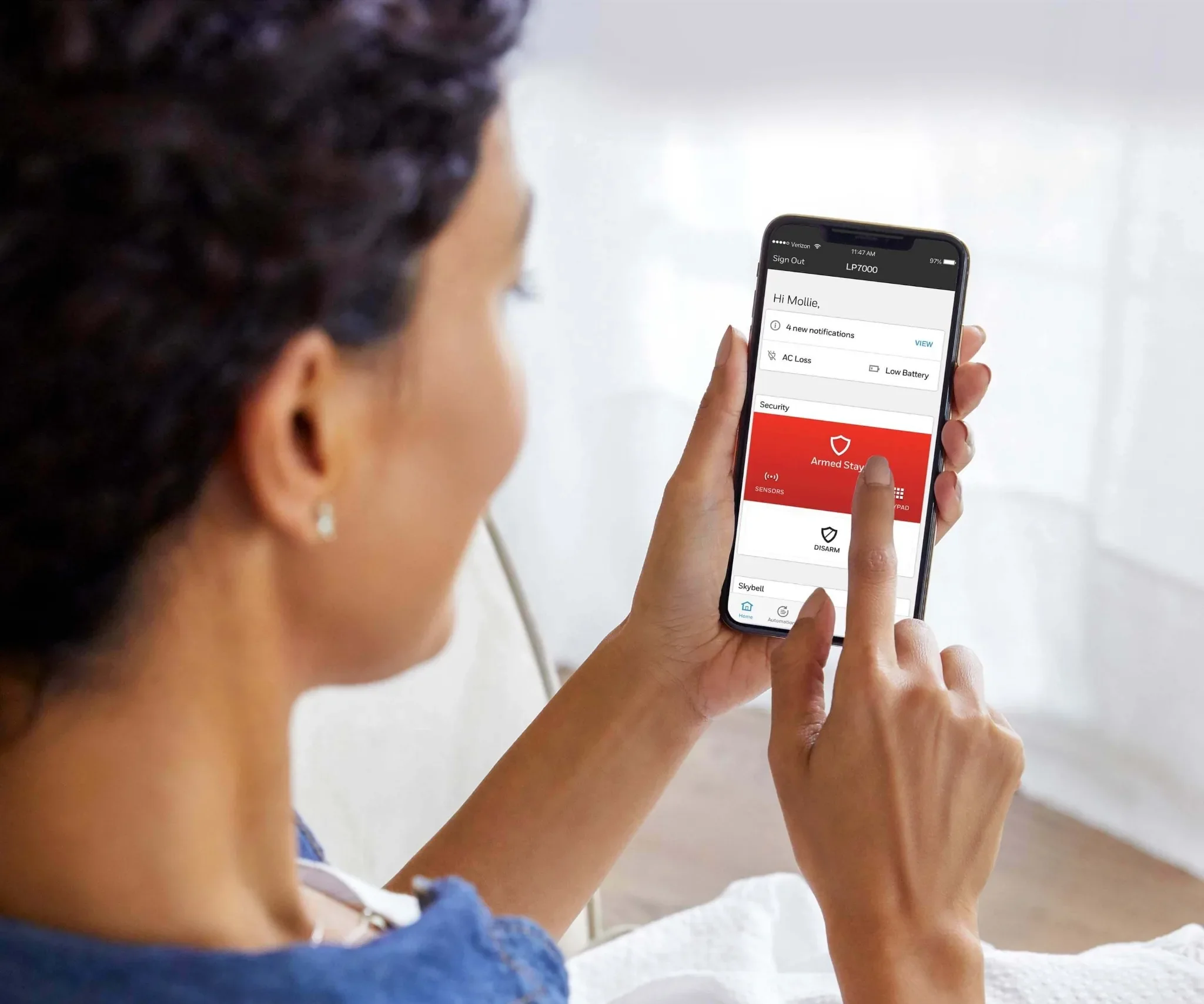 A person with curly dark hair holding a smartphone displaying a security alarm app with a red armed stay alert message