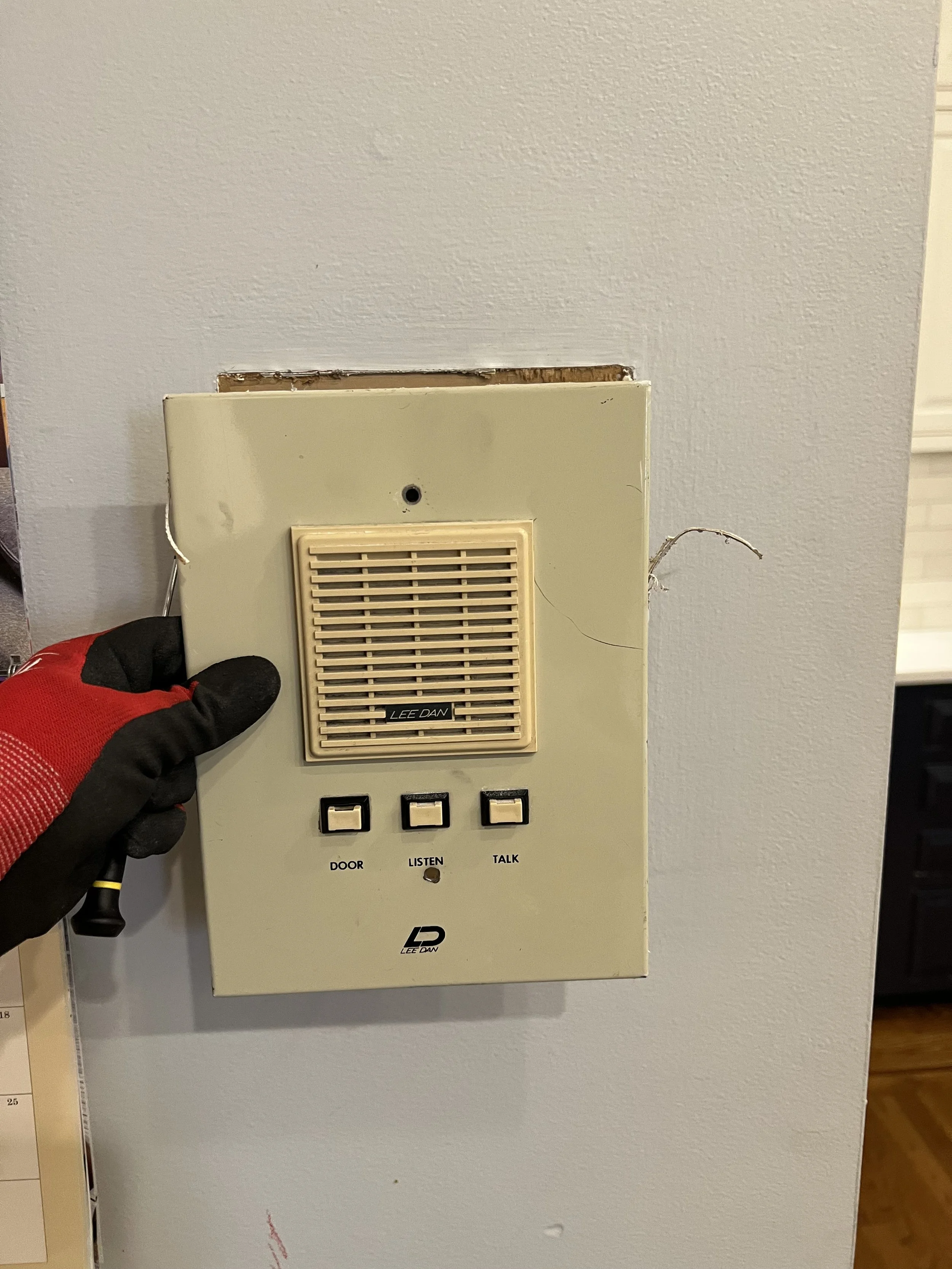 An old intercom or door entry system on a wall with a person's gloved hand holding a small black antenna or remote near it. The device has a speaker grille, three buttons labeled 'DOOR,' 'LISTEN,' and 'TALK,' and a small hole at the bottom. The surface of the device shows some wear and tear.