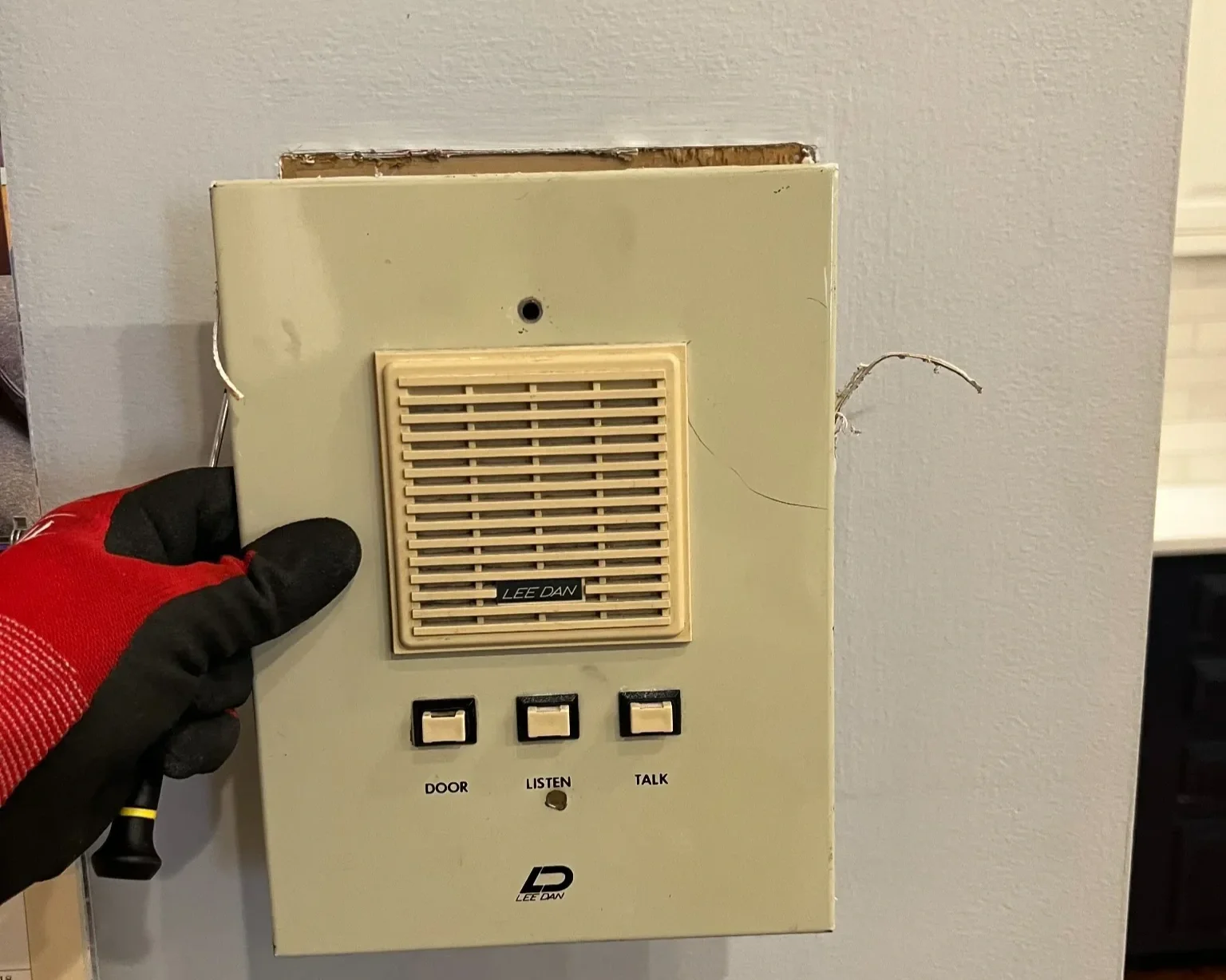 An old beige intercom system with a speaker, three buttons labeled 'DOOR', 'LISTEN', and 'TALK', and a person's gloved hand holding a screwdriver next to it.