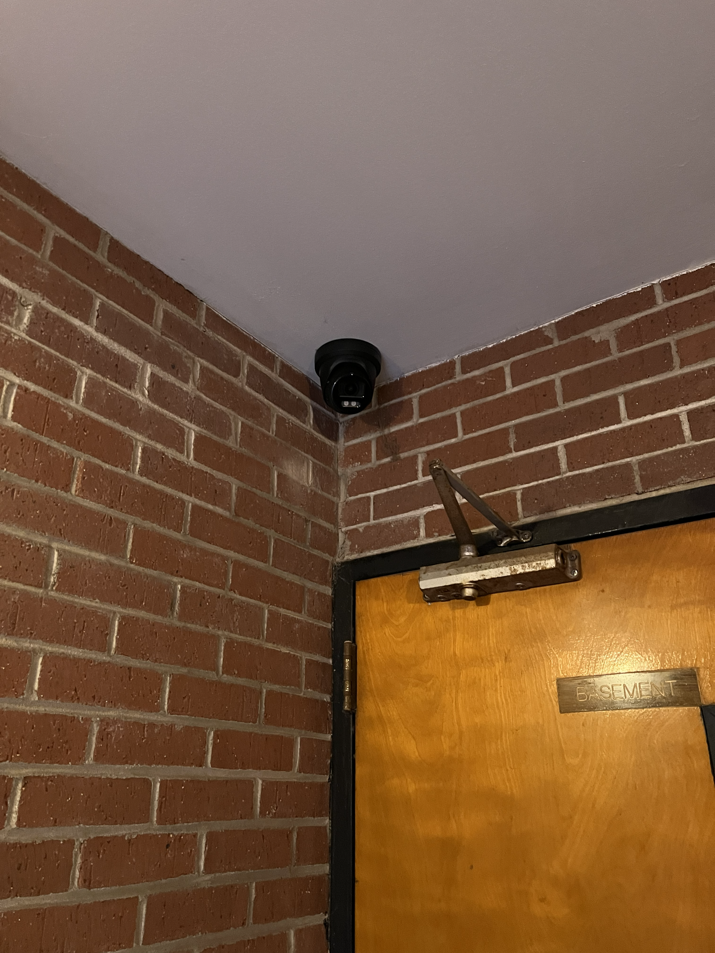 Security camera installed in the corner ceiling near a wooden door labeled "Basement" in a brick-walled room.