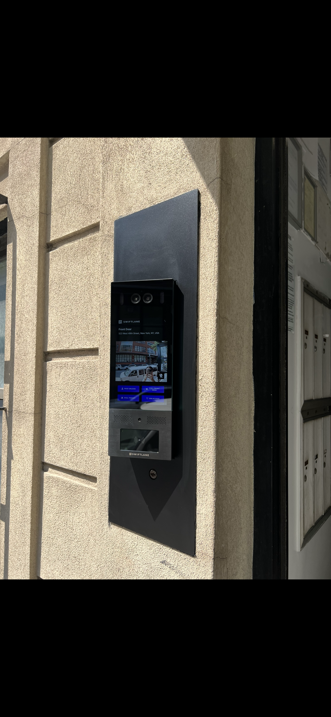 A modern intercom and video door entry system mounted on a beige exterior wall, with a screen displaying a street view and options for door access.