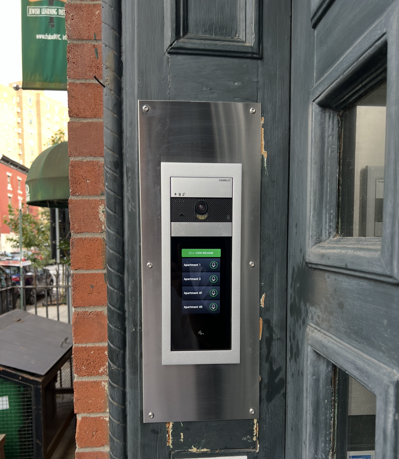 Intercom system with a camera and a touchscreen display showing apartment options, mounted on a brick and wooden wall outside a building.