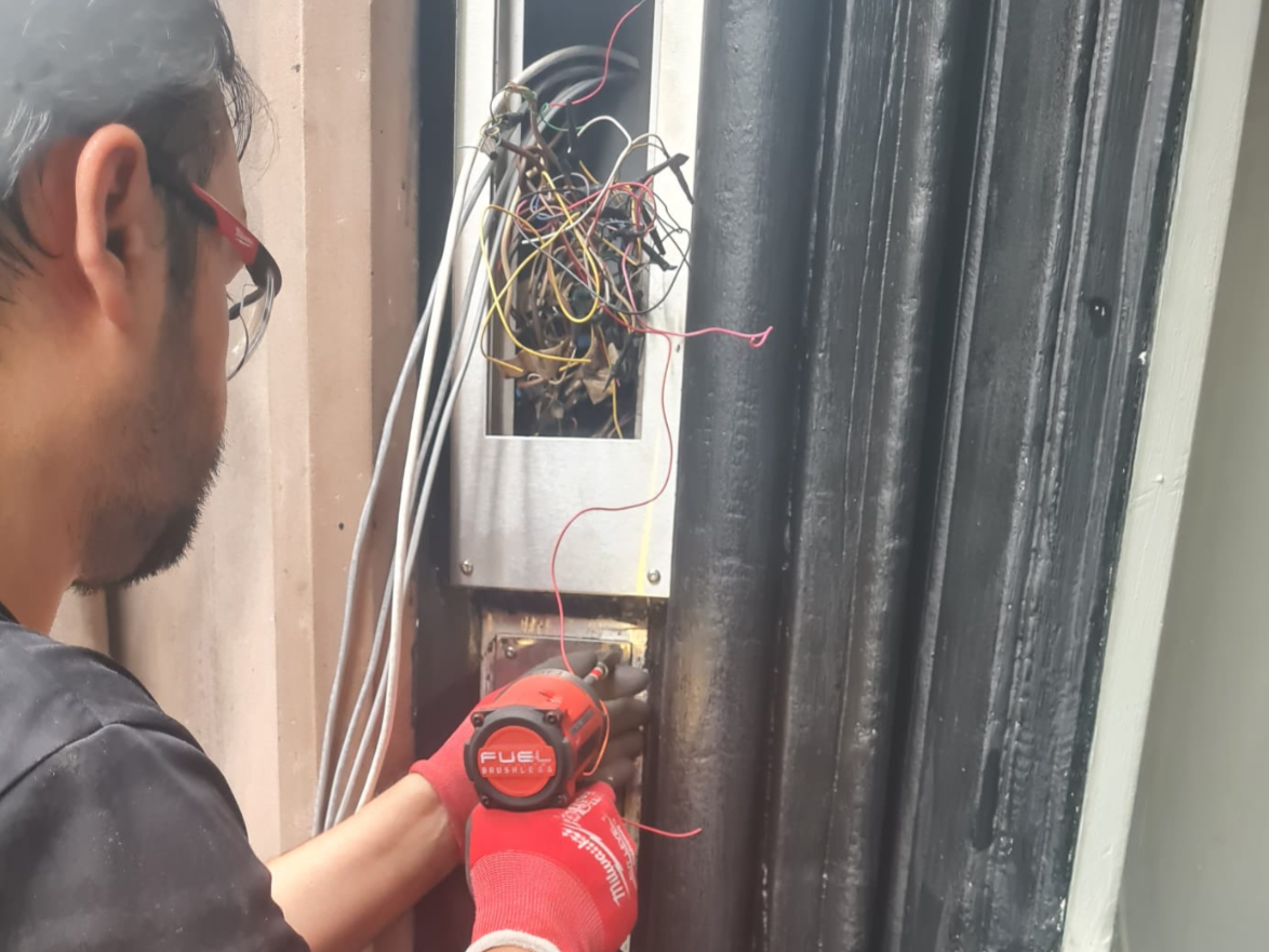 Person working on an electrical panel with exposed wiring, wearing red gloves and using a cordless drill.