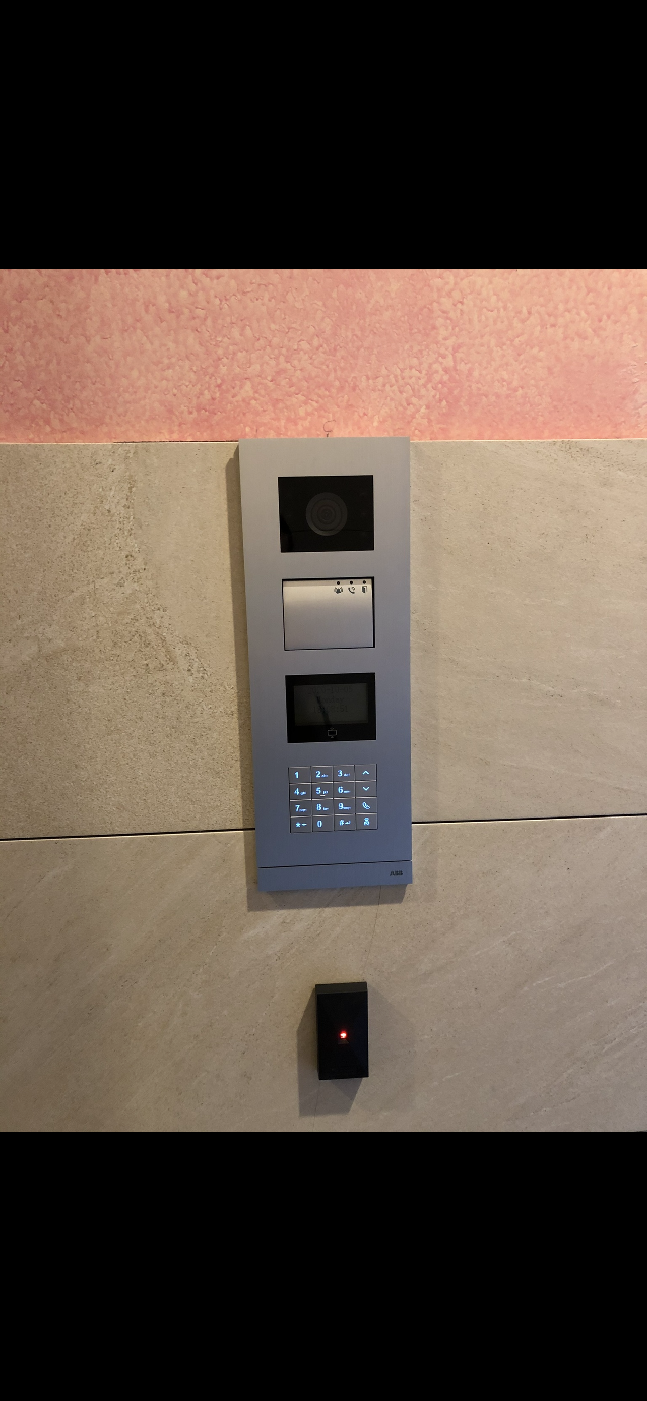 A modern intercom system mounted on a wall with a keypad and video screen, and a small black sensor below.