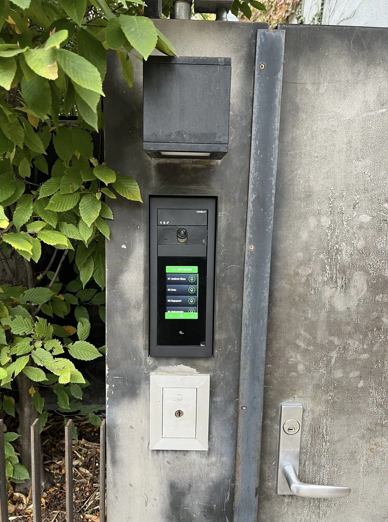 Video intercom and access control panel mounted on a concrete wall near a door with a silver handle, surrounded by green foliage.