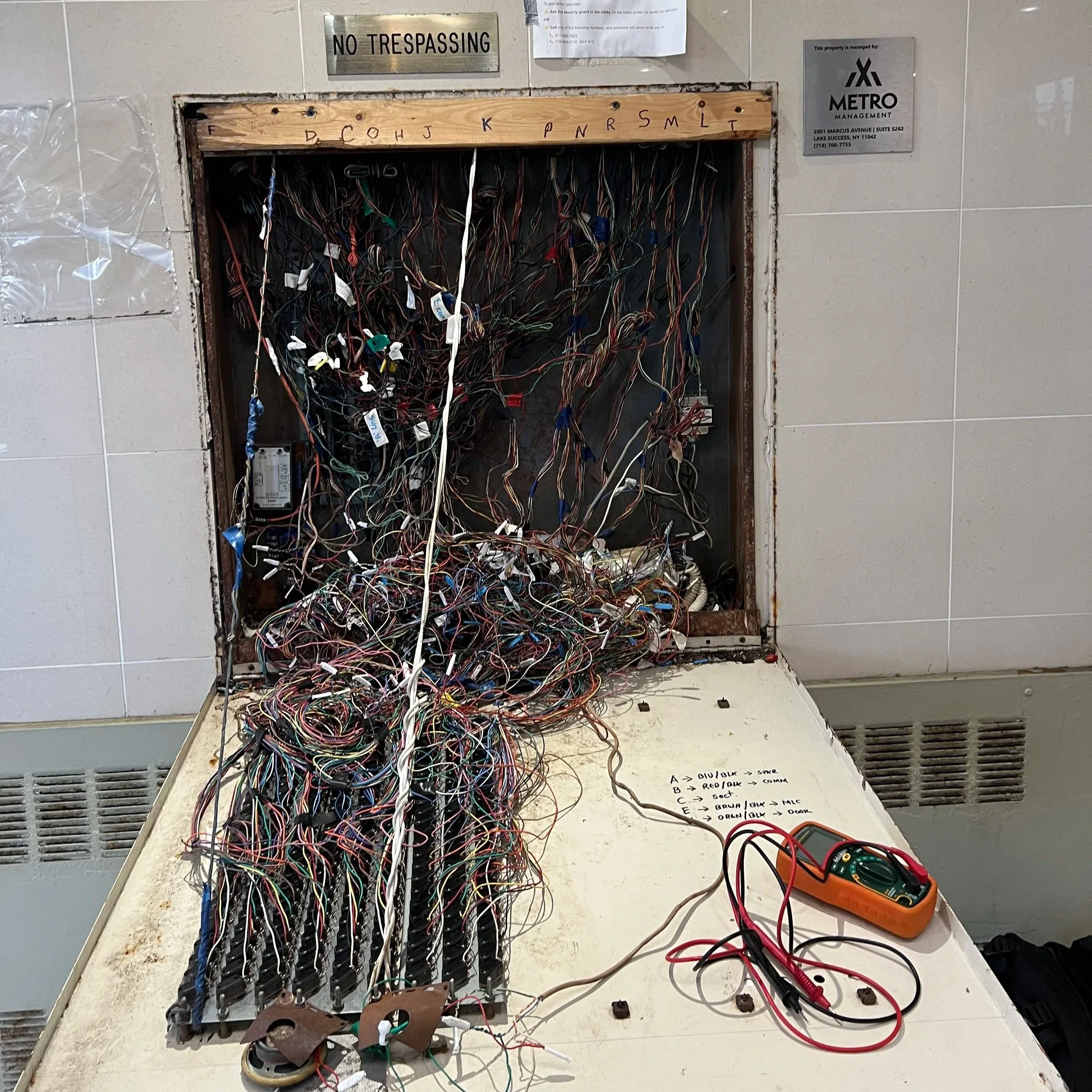 Open electrical panel with a large mass of tangled multicolored wires, some with labels, and a multimeter with red and black probes connected on the right side. There is a handwritten note with wire color codes and functions on the panel's bottom right.