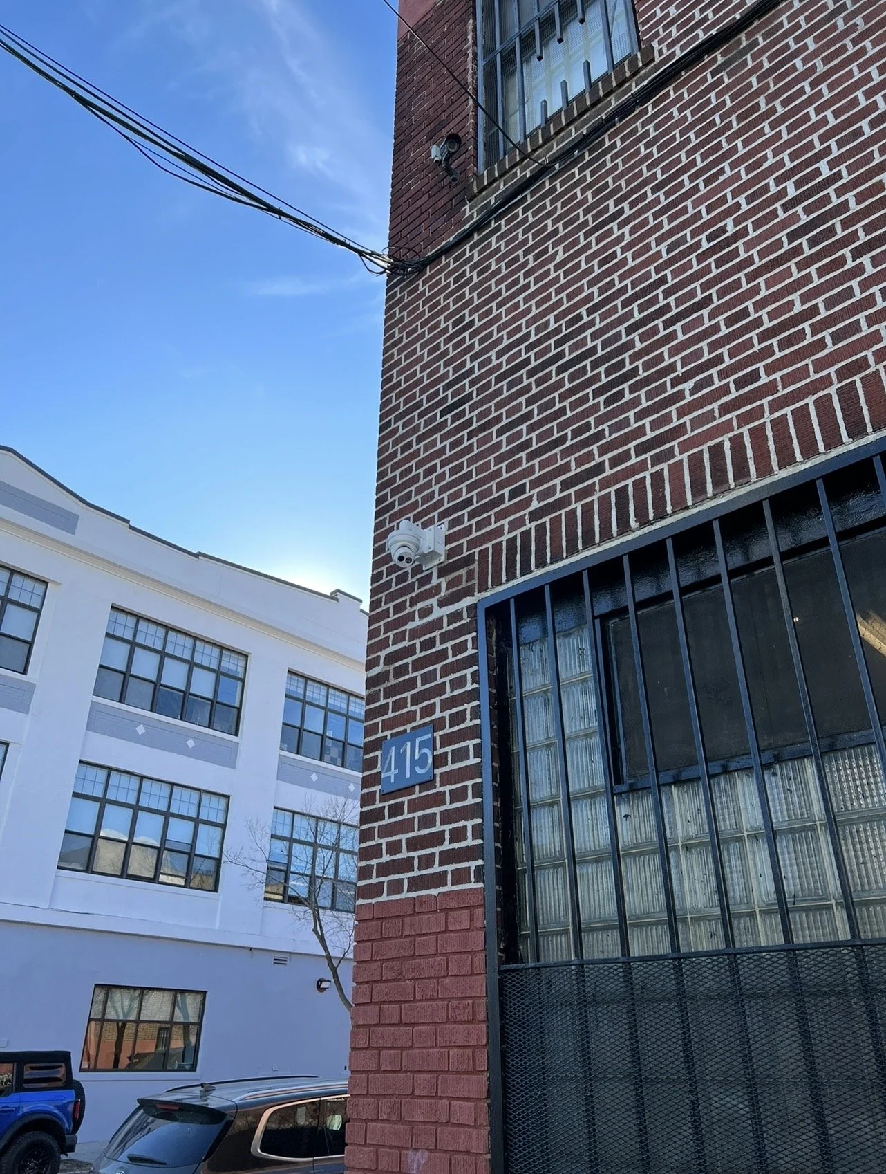 A brick building with a blue address plaque displaying '415', a security camera, and barred windows. In the background is a white building with large windows under a clear blue sky.