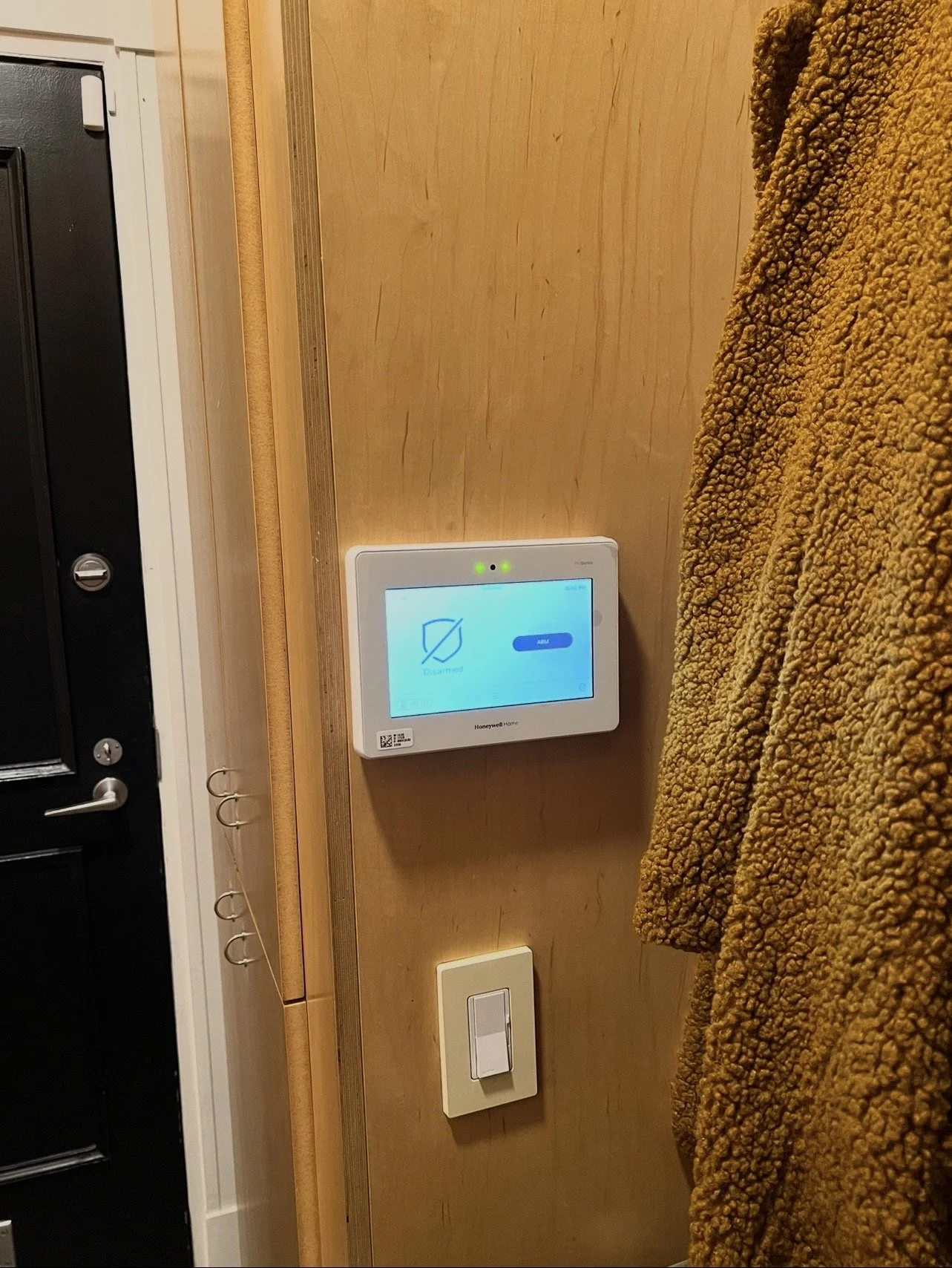 Smart home security control panel on a wooden wall, with a brown textured blanket partially visible on the right, and a black door with a lock on the left.