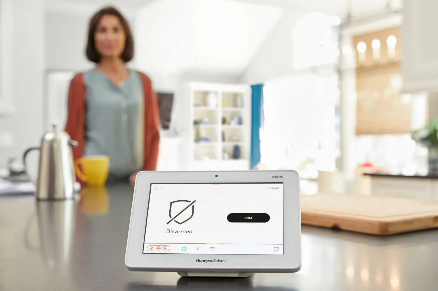 Smart home security control panel displaying 'Disarmed' and 'ARM' with a blurred woman in the background in a kitchen.
