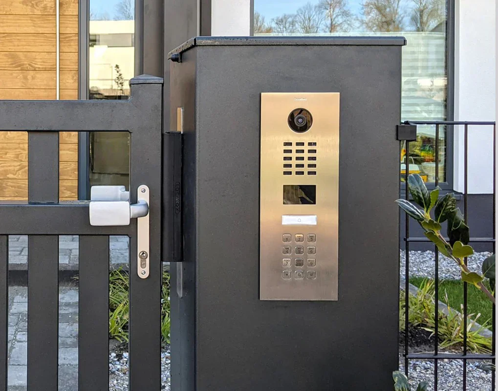 Front view of a black gate with a built-in modern intercom system, including camera, keypad, and speaker, outside a residential building.