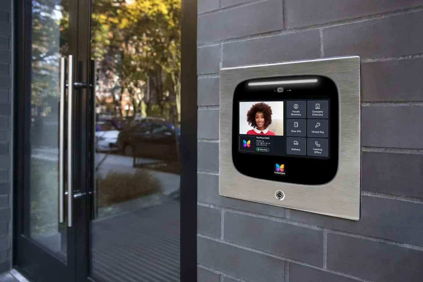 A modern intercom panel with a touchscreen at the entrance of a building, displaying a smiling woman and various options for access control and directory services, mounted on a brick wall outside.