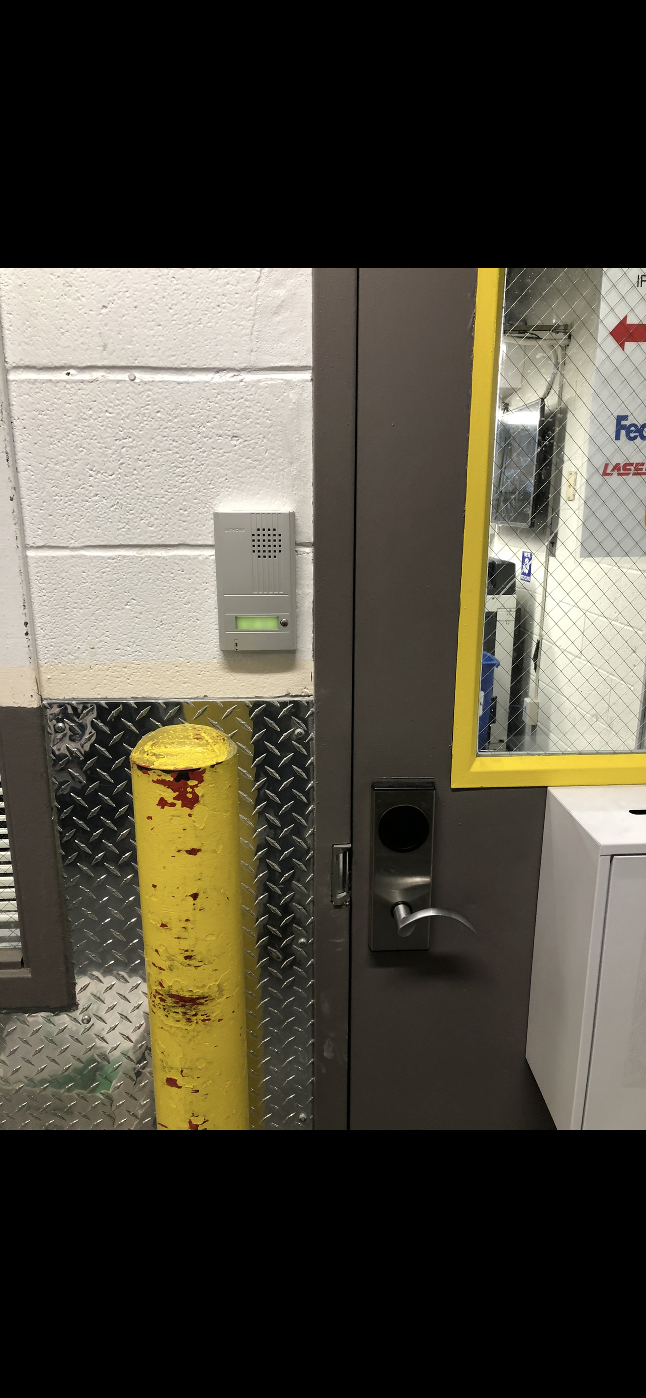 Public door entrance with a yellow bullet pole, an intercom system on a white cinder block wall, and a glass door with a push bar.