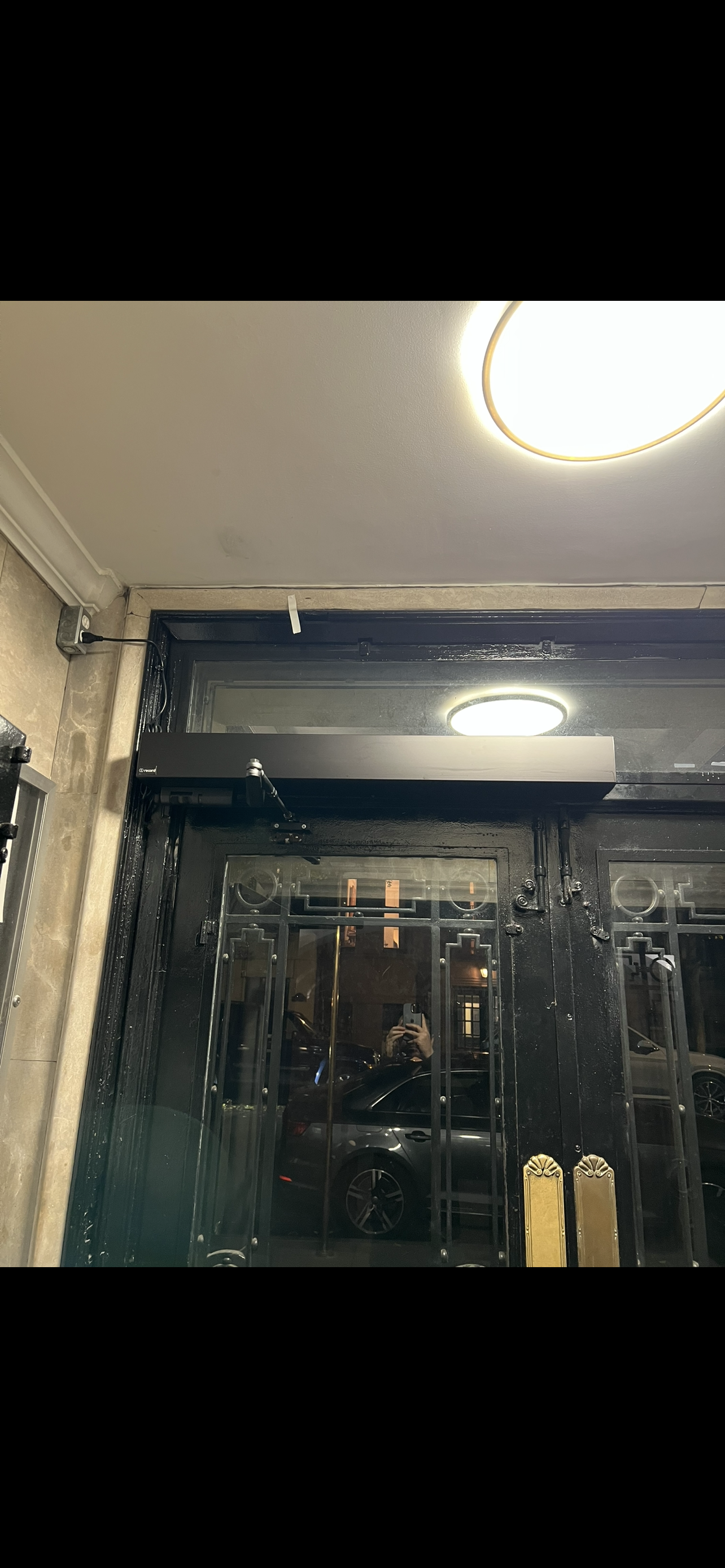 Photo of a black glass door with gold handle, seen from inside a building at night. Reflection of a car and the photographer with a phone is visible in the glass.