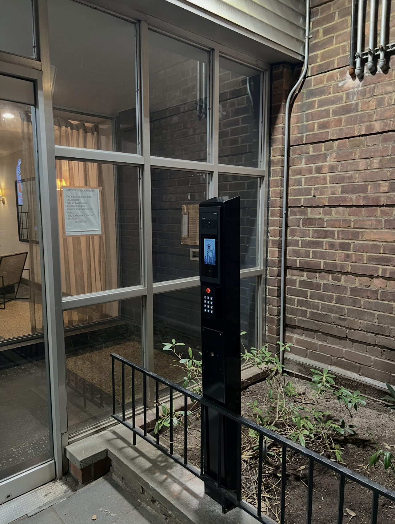 A modern digital intercom and keypad access system installed outside a building, next to a glass entry door and small garden with plants, brick wall, and window.